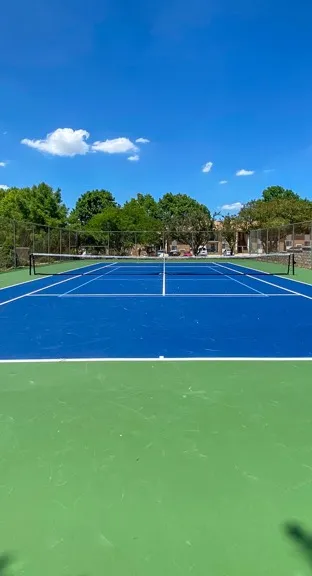 A bright, empty tennis court under a clear blue sky with fluffy clouds. Green and blue surfaces are surrounded by trees and buildings, creating a peaceful setting.
