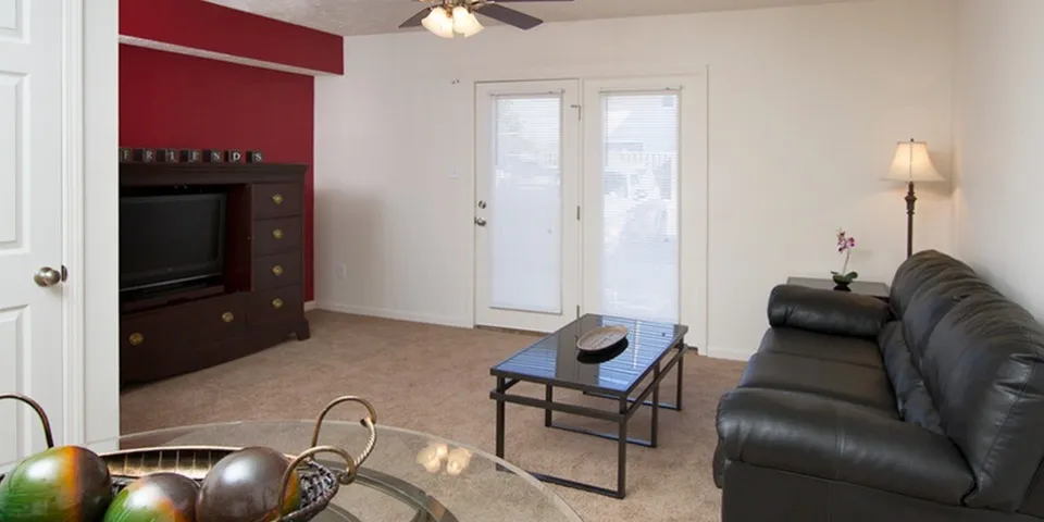 Furnished living room with carpeted floors, neutral walls, a bold red accent wall, and double French doors leading to a private outdoor space—ideal for relaxing or entertaining.