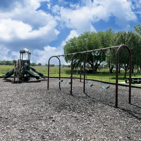 Outdoor play area with multiple swings and a jungle gym set on mulch with scenic views of open green space and mature trees—a family-friendly amenity for residents.