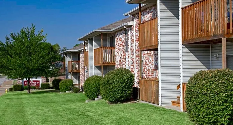 Row of two-story buildings at Grandview Village featuring red brick and light siding, wooden balconies, and lush green lawns with trimmed bushes.