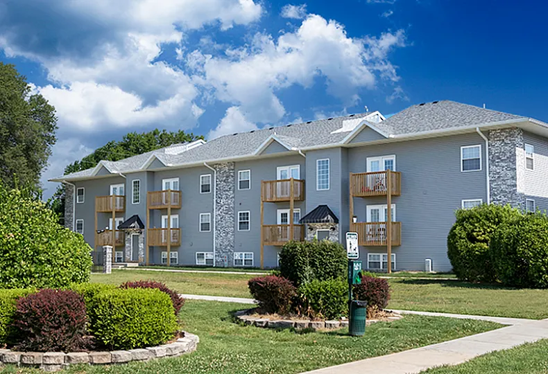 A three-story apartment building with gray siding, stone accents, and wooden balconies, surrounded by lush green landscaping and a neatly trimmed lawn. A walking path leads to the entrance, and a pet waste station is visible in the foregrou