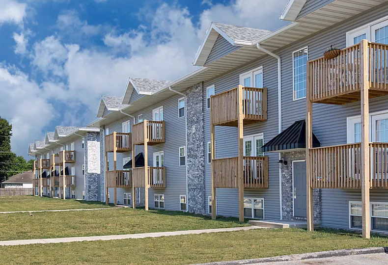A side view of a row of three-story apartment buildings at Concord Manor, featuring gray siding, brick accents, and wooden balconies. The well-maintained lawn and paved walkways run parallel to the buildings, with a clear blue sky and trees