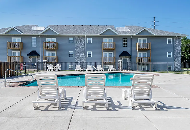 A three-story apartment building at Concord Manor with gray siding, brick accents, and wooden balconies. In the foreground, a fenced-in outdoor pool area features three white lounge chairs facing the water. Additional chairs and tables are