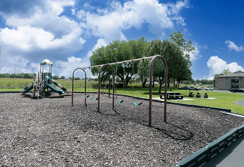 A playground with a swing set and a large play structure featuring multiple slides and climbing areas, surrounded by mulch ground covering. In the background, there are picnic tables on a grassy area with trees providing shade. A parking lo