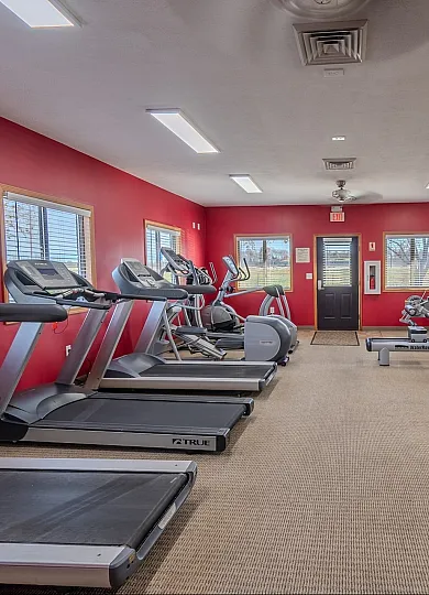 A well-lit fitness center at Concord Manor Apartments in Springfield, MO, featuring red walls and large mirrors. The gym is equipped with treadmills, ellipticals, stationary bikes, and strength training machines, all neatly arranged. Window