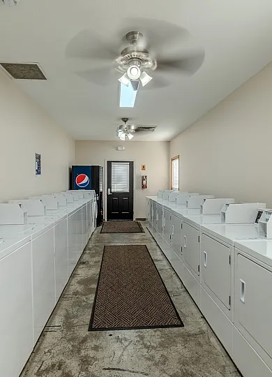 A community laundry room featuring rows of white top-loading washing machines and dryers on both sides. The space has beige walls, a concrete floor with two brown runner rugs, and ceiling fans with lights. A black door with a window at the