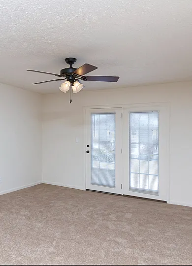 A spacious living room with beige carpeting and off-white walls. A black ceiling fan with four wooden blades and three attached light fixtures is centrally mounted on the ceiling. A set of white French doors with built-in blinds leads to an