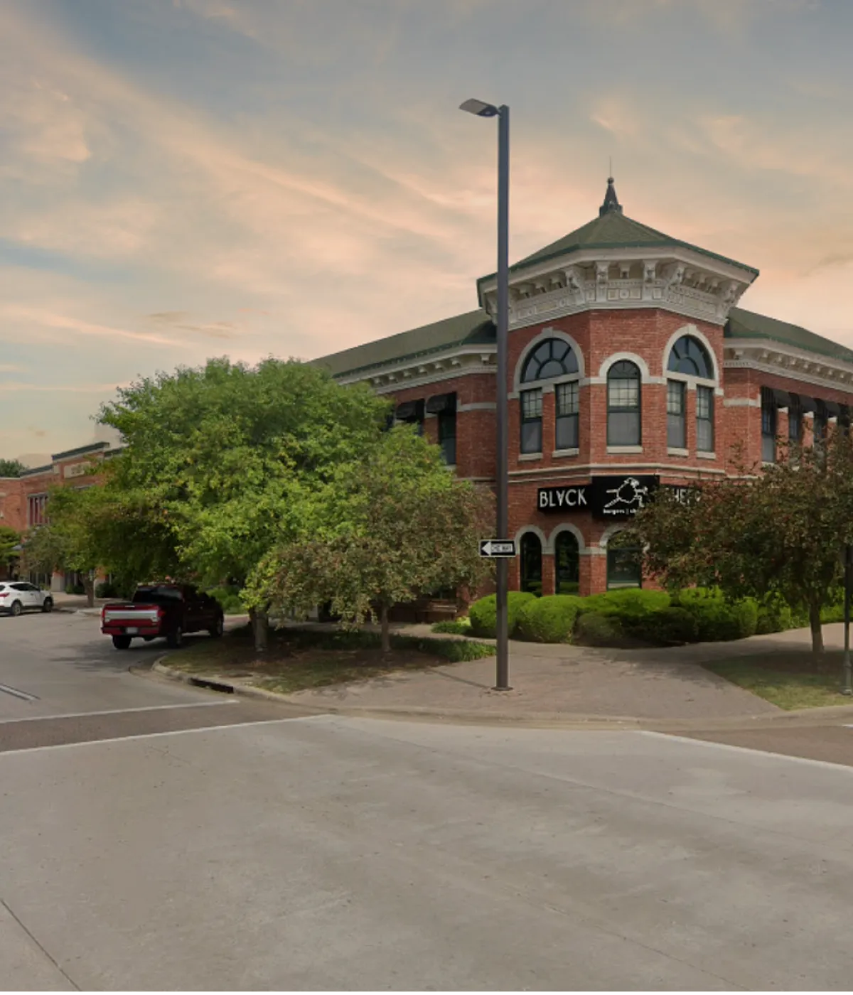 A two-story historic red brick building with ornate white stone detailing, large arched windows, and a green-roofed turret stands at a corner under a soft, sunset sky. A business sign reading 'BLYCK SHEEP' is mounted between the f