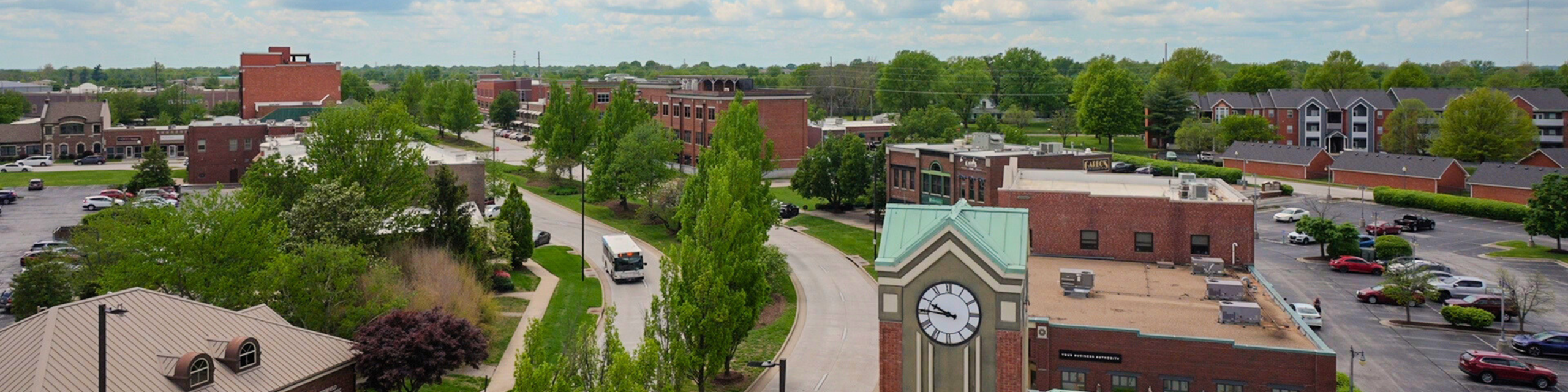 Aerial view of Chesterfield Village in Springfield, Missouri, showcasing a tree-lined street, clock tower, brick office buildings, apartment complexes, and parking areas on a partly cloudy spring day.