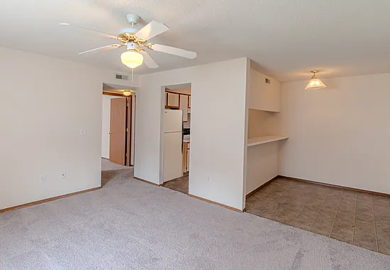 View of an upgraded apartment unit at Huntley Ridge East in Springfield, MO, featuring a partial open kitchen with white appliances and light wood cabinetry, a breakfast bar, tile flooring in the kitchen, and carpeted living area with a cei