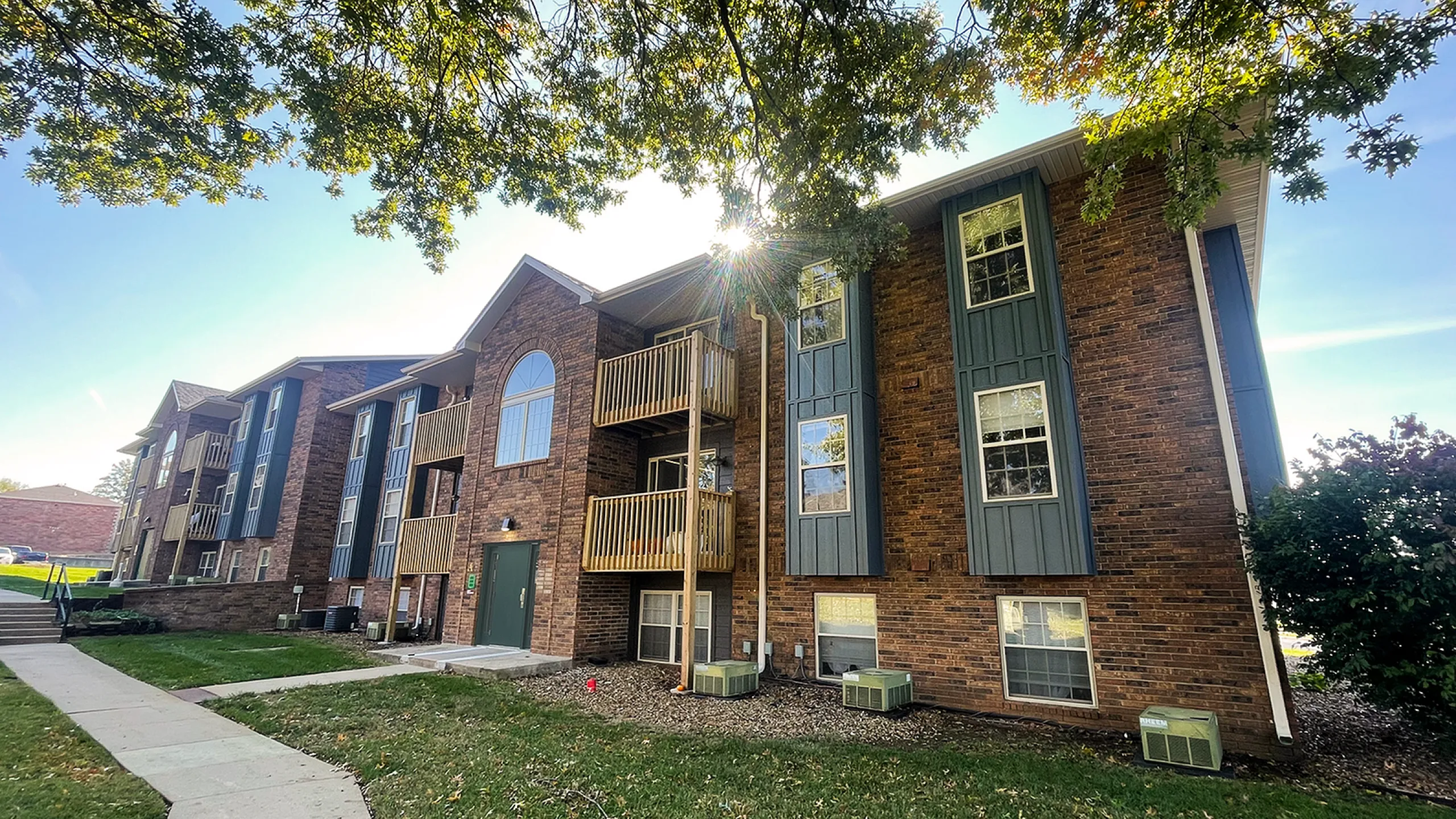 Charming brick apartment building surrounded by lush greenery under a clear blue sky, featuring spacious balconies and large windows.