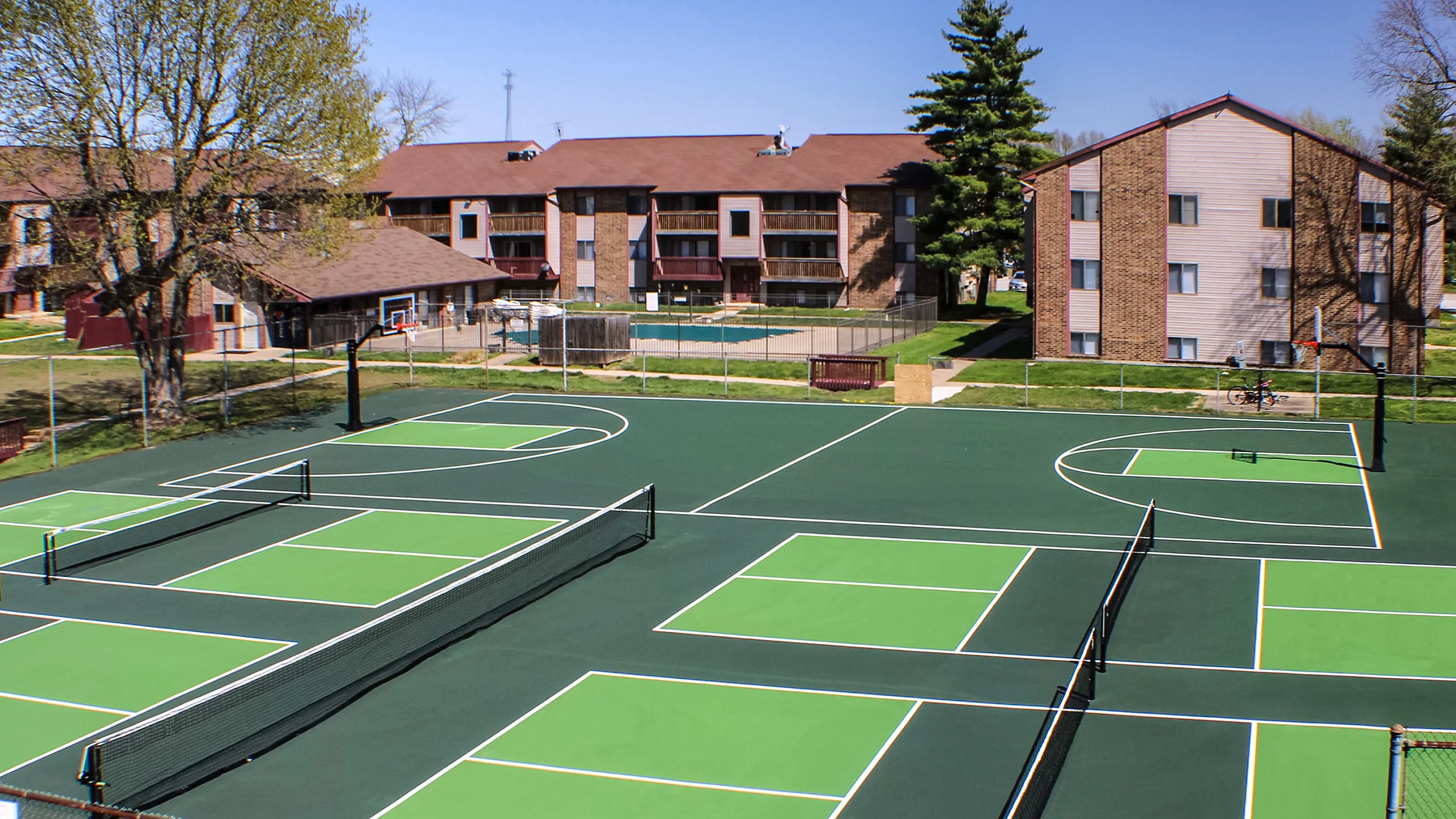 Charming apartment complex with modern tennis and basketball courts, surrounded by lush greenery and brick buildings.