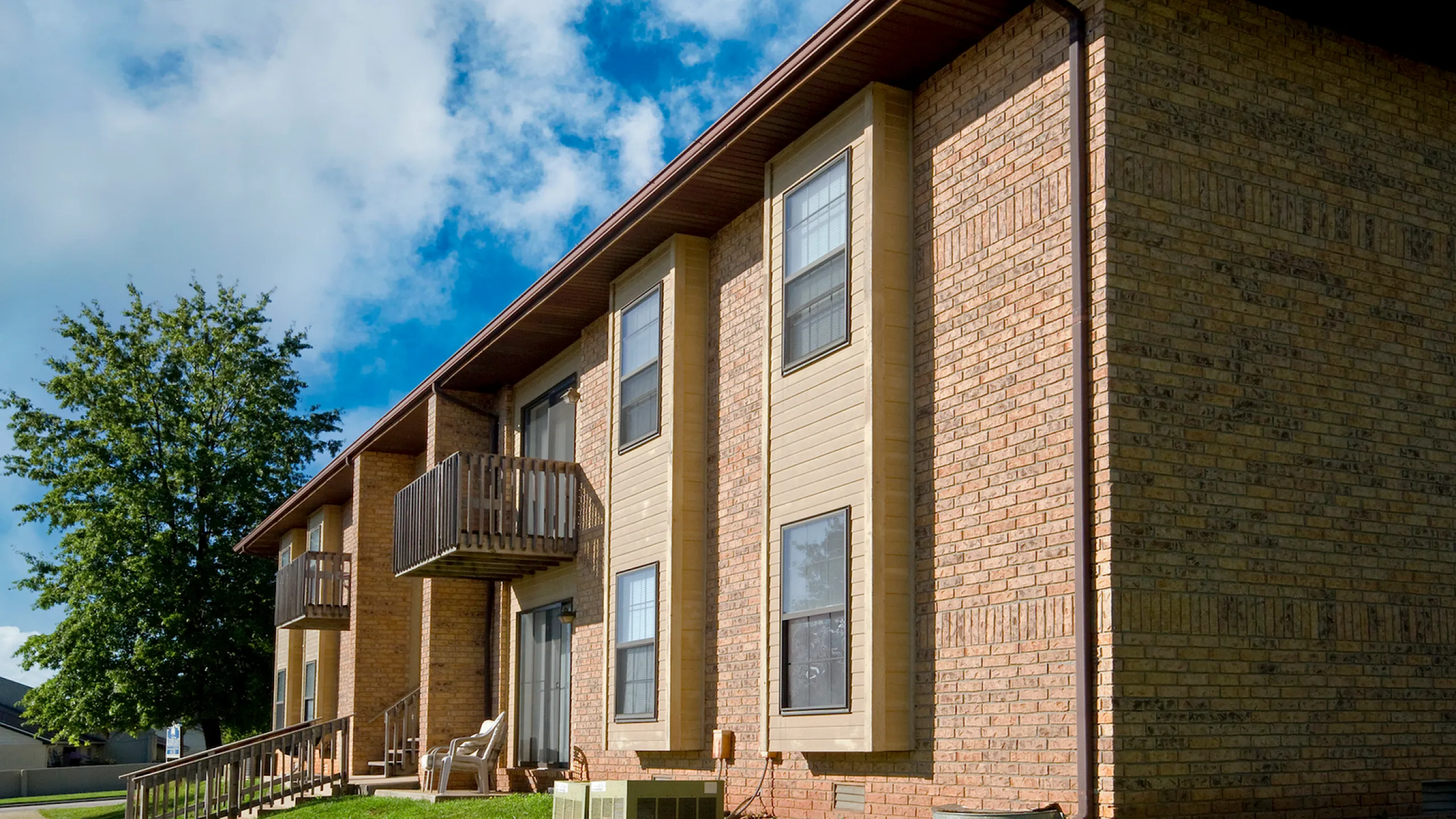 Charming brick apartment complex with spacious balconies, lush green lawn, and bright surroundings under a clear blue sky.
