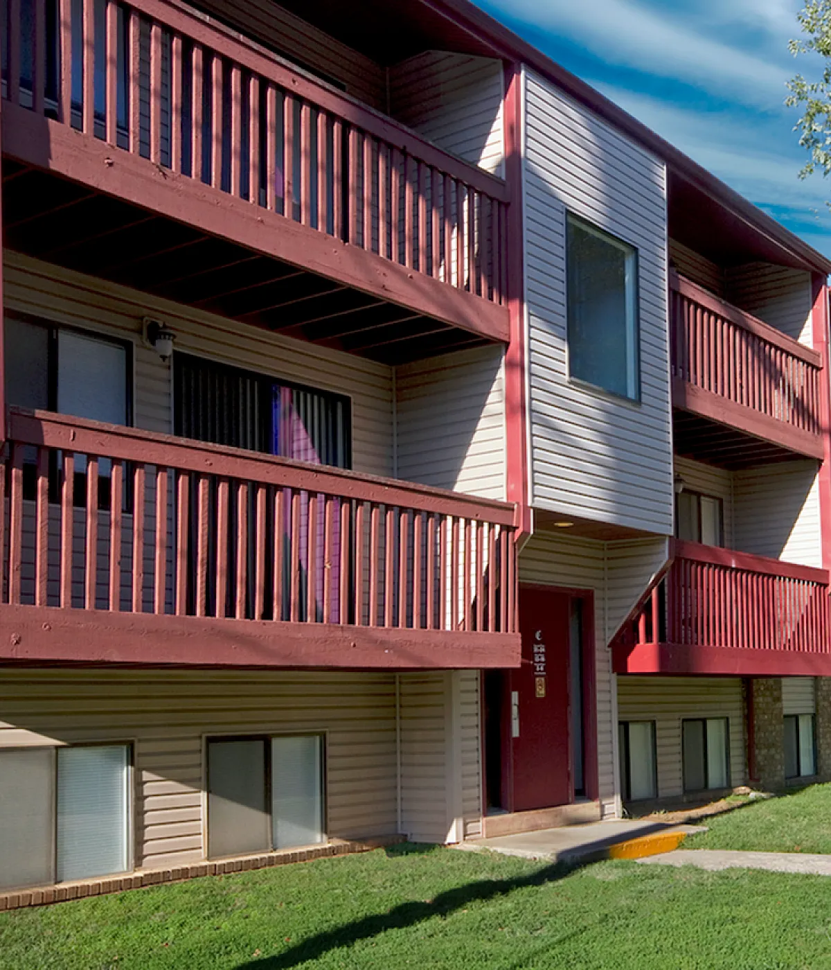 Exterior view of Briarwood Village Apartments in Springfield, Missouri, showing a two-story building with beige siding, brick accents, and maroon patios and balconies. A large mature tree provides shade over the neatly maintained lawn and s