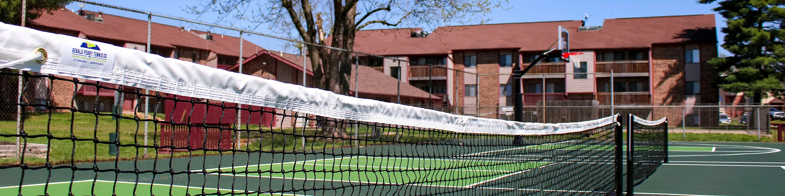 Vibrant tennis court with well-maintained green surface surrounded by lush trees and modern apartment buildings under a clear blue sky.