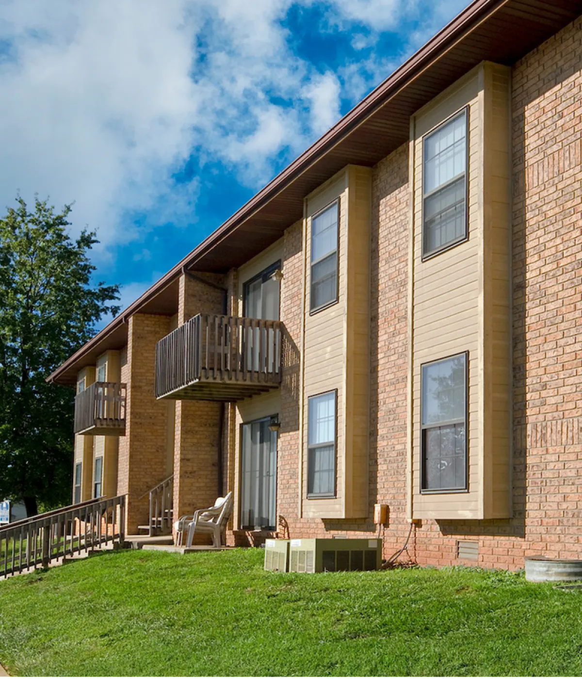 Charming brick apartment building with private balconies, sunlit windows, and lush green surroundings on a bright day.