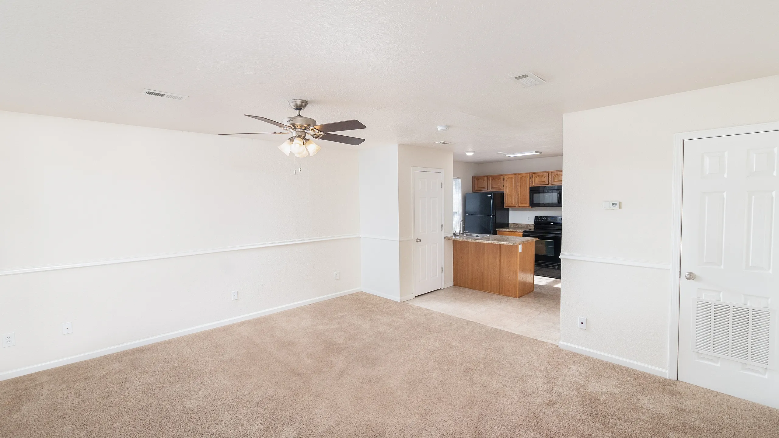 Bright and open living space with plush carpeting, modern ceiling fan, and an inviting kitchen with warm wood cabinetry.