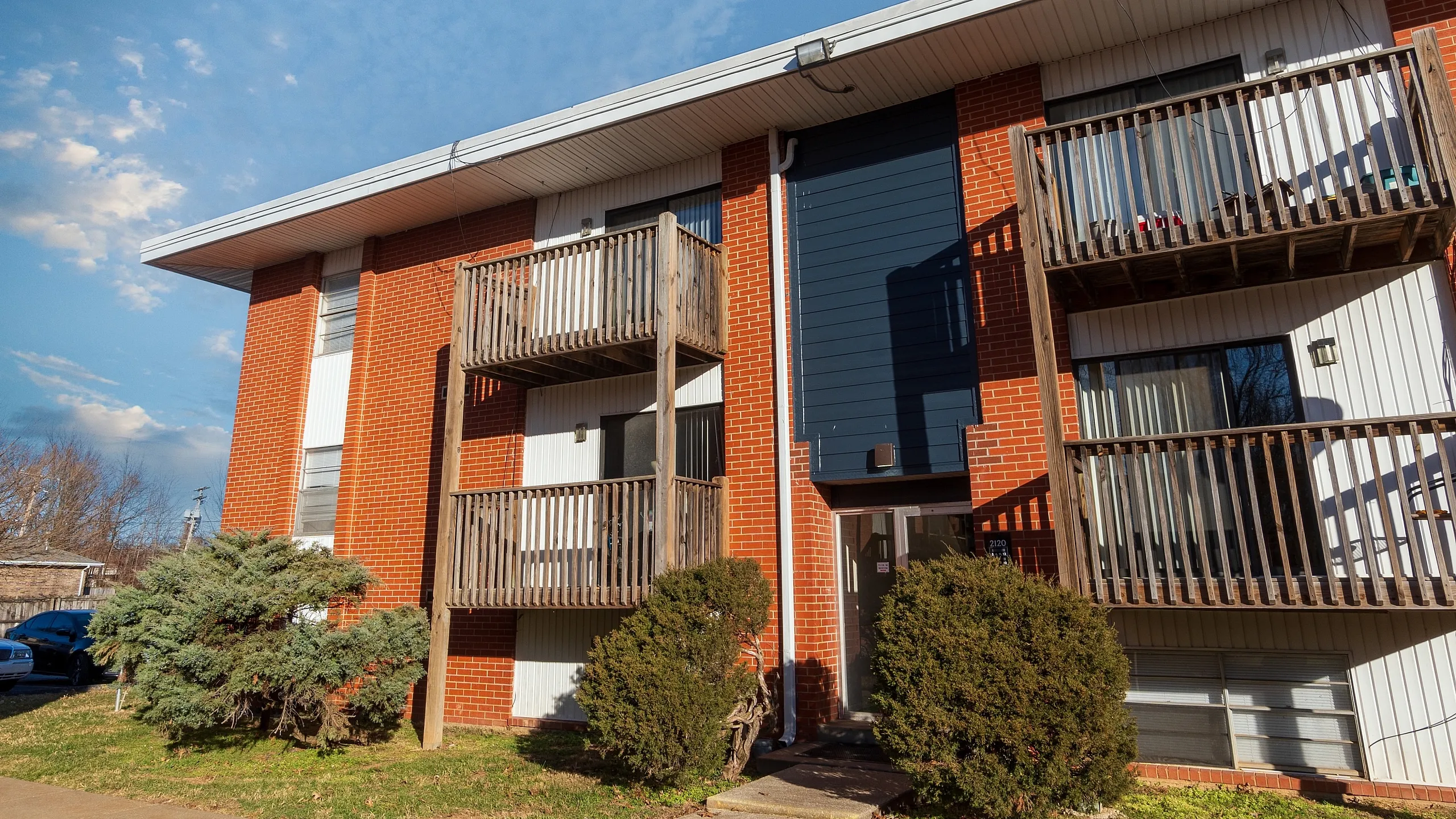 Front exterior of a Seminole North Apartments building with brick fa&ccedil;ade, vertical blue accent panel, stacked wooden balconies, ground-level entry, and trimmed shrubs along the walkway.