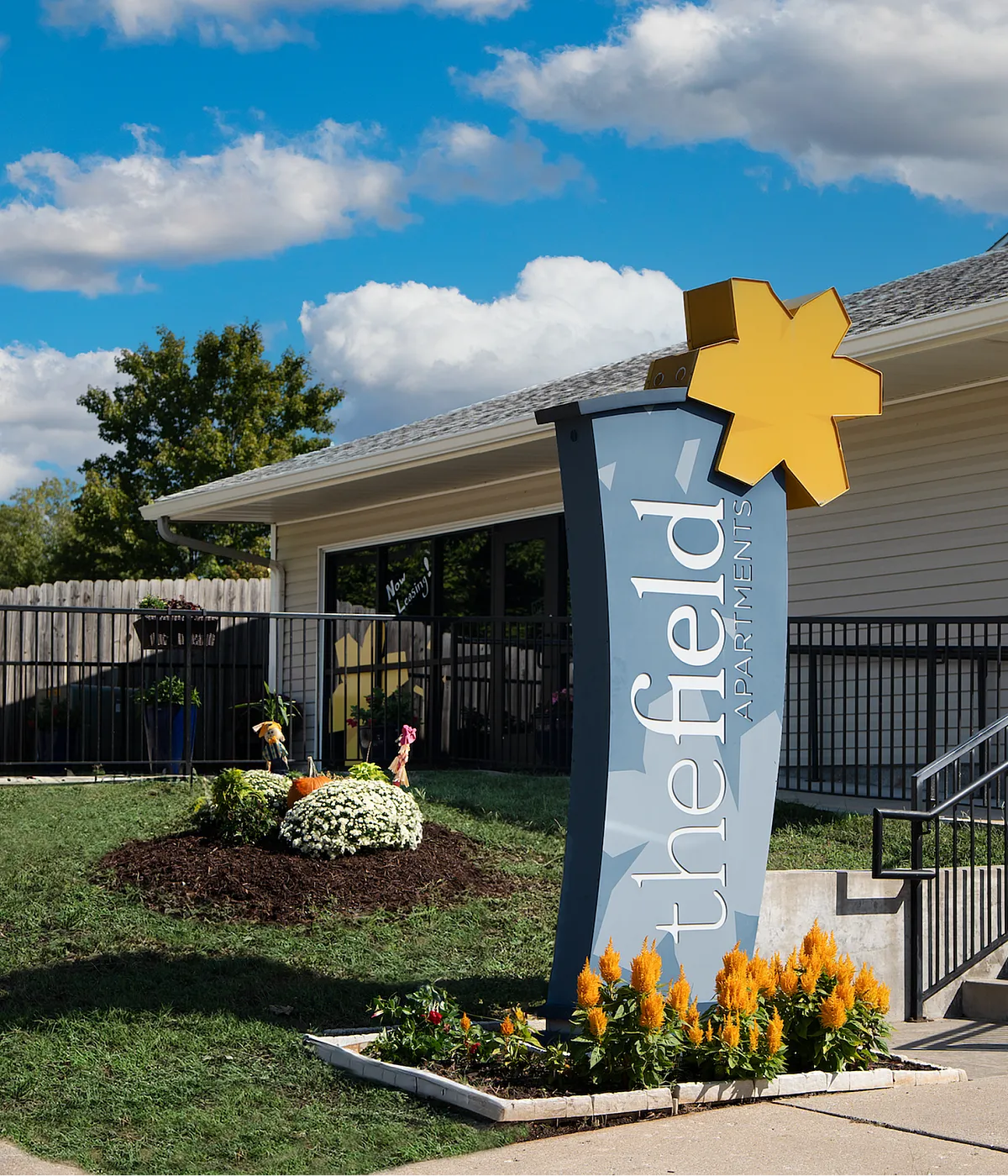 Bright and welcoming entrance to The Field Apartments, highlighted by vibrant landscaping and modern signage under a clear blue sky.