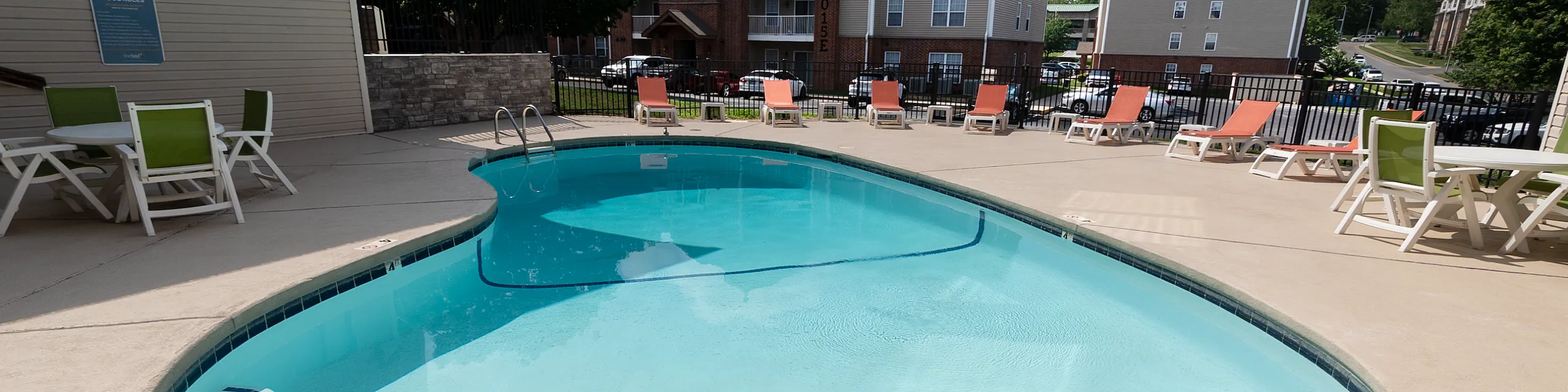 Inviting apartment complex with a crystal-clear outdoor swimming pool, surrounded by sun loungers and vibrant green chairs, perfect for relaxation under bright blue skies.
