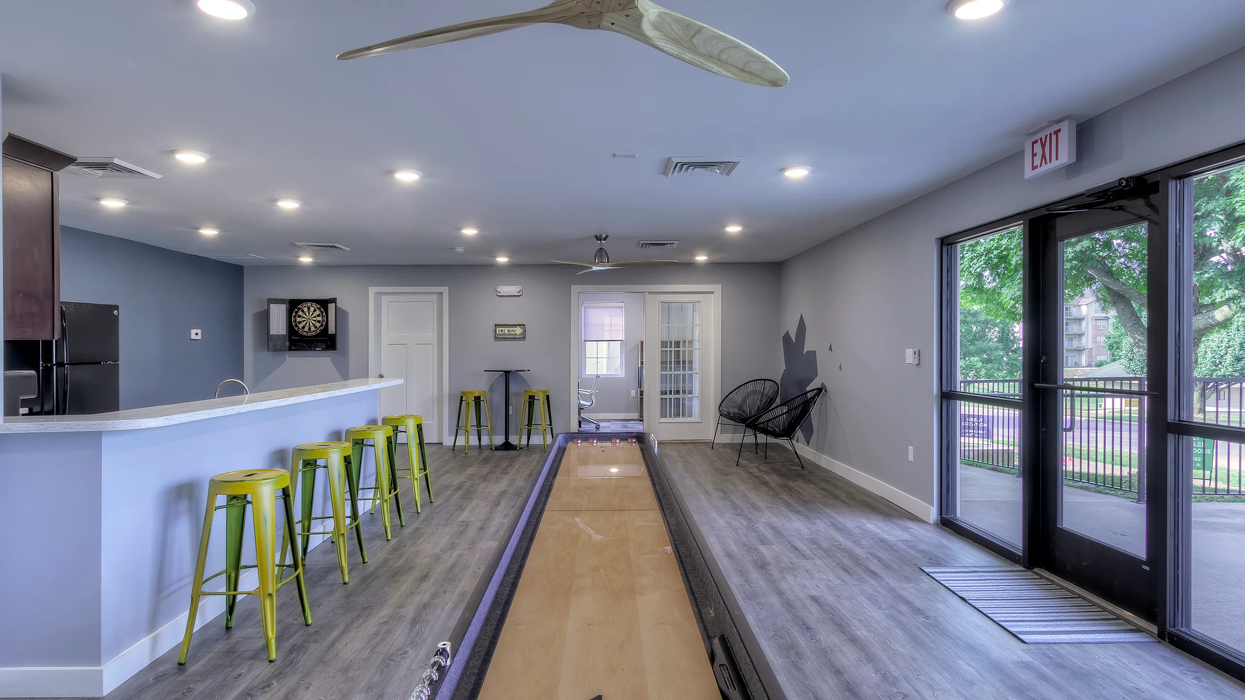 Community lounge with a shuffleboard table centered in the room, bar seating with green stools along a kitchen counter, recessed ceiling lights and modern ceiling fans, gray wood-style flooring, and glass doors leading to an outdoor patio.