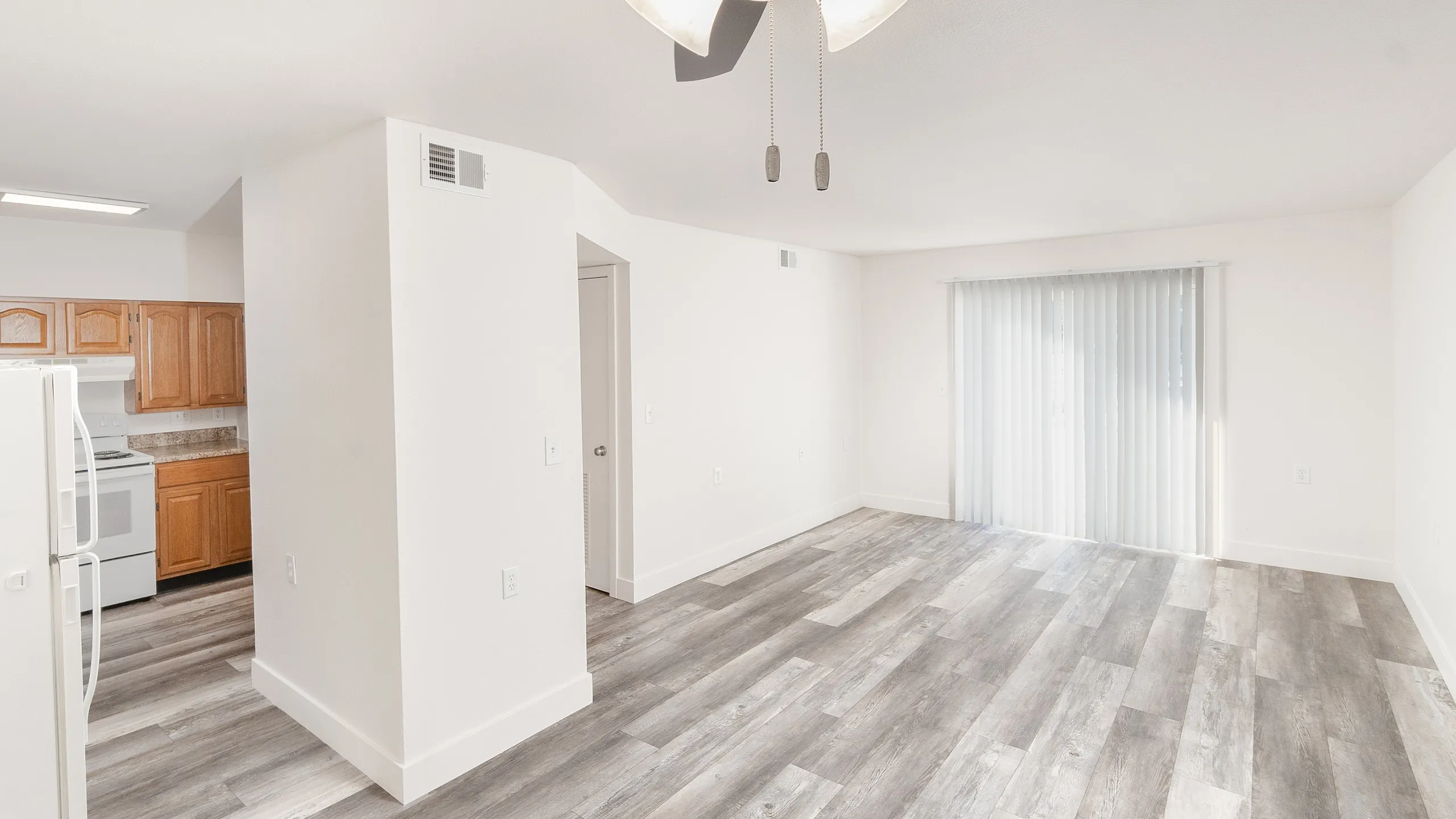Bright and airy living space with modern ceiling fan, open floor plan featuring elegant wood-look flooring, cozy kitchen with wooden cabinets, and large sliding glass doors inviting abundant natural light.