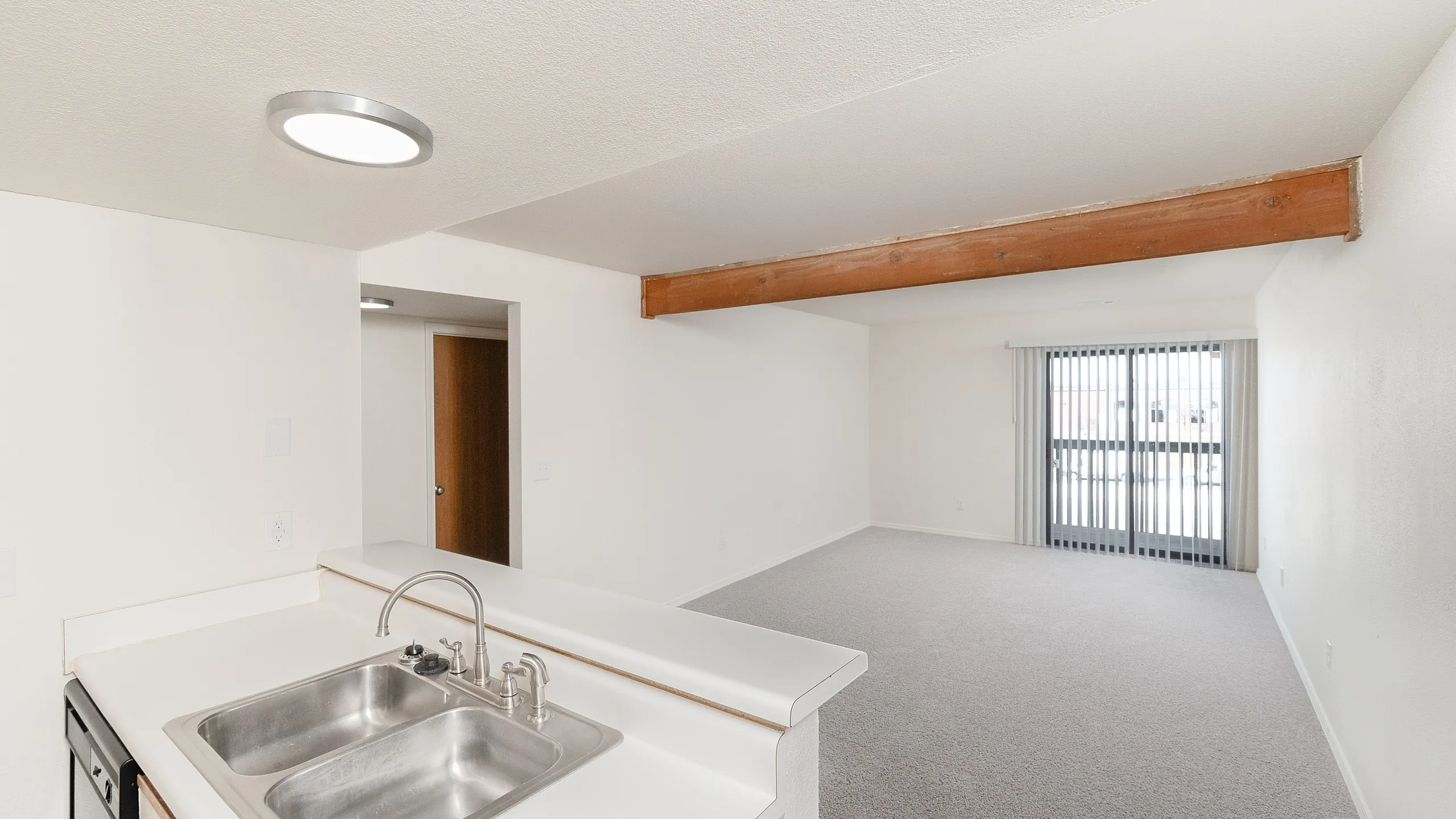 Living room with open kitchen pass-through, neutral carpet, ceiling beam detail, sliding patio door with vertical blinds