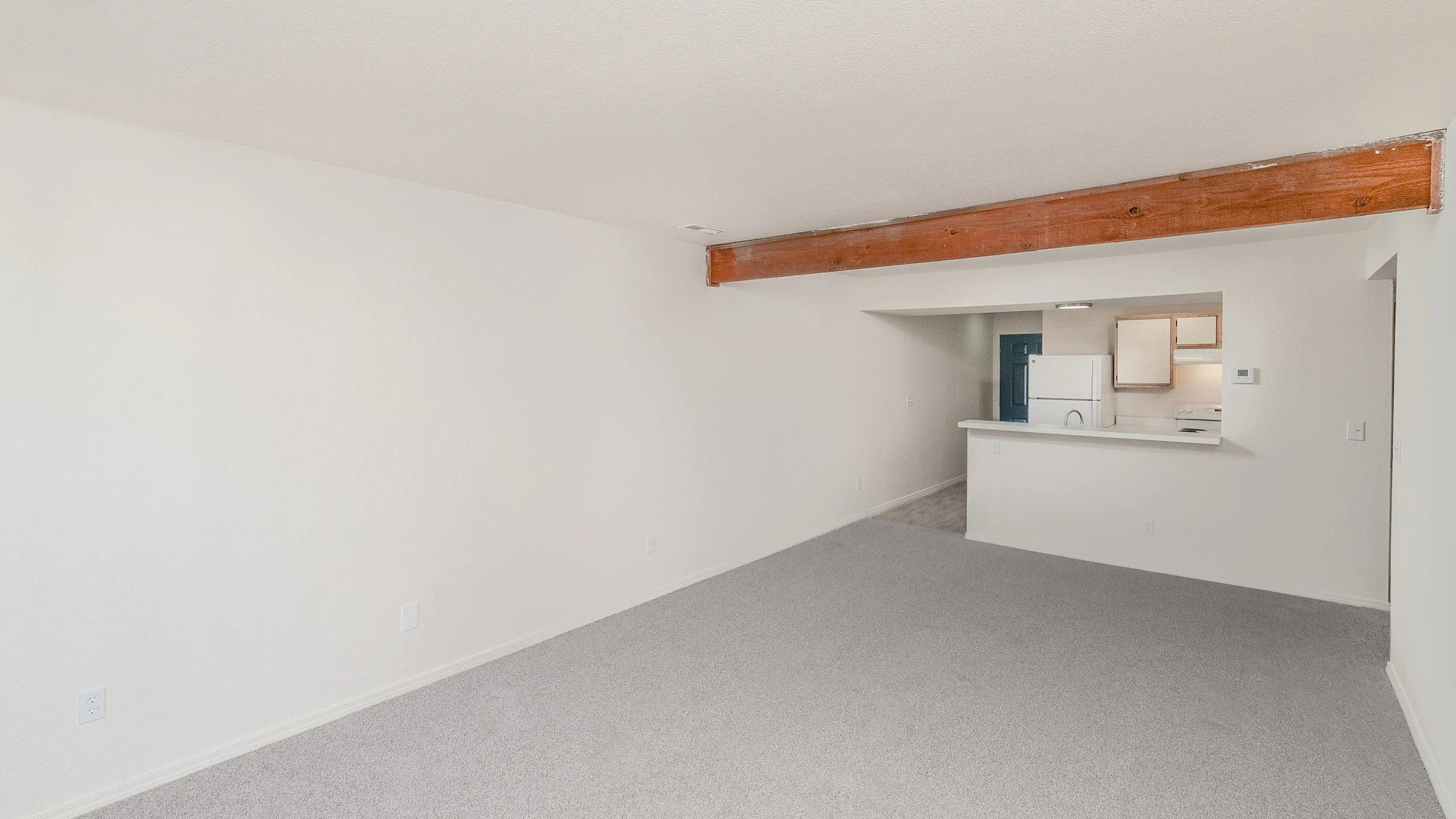 Spacious living room with modern open-concept kitchen, featuring exposed wooden beams and plush carpet flooring.