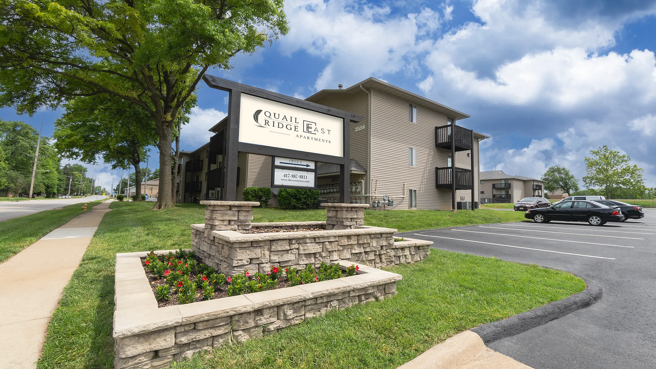 Inviting view of Quail Ridge East Apartments with well-maintained landscaping, bright signage, and ample parking under a sunny blue sky.