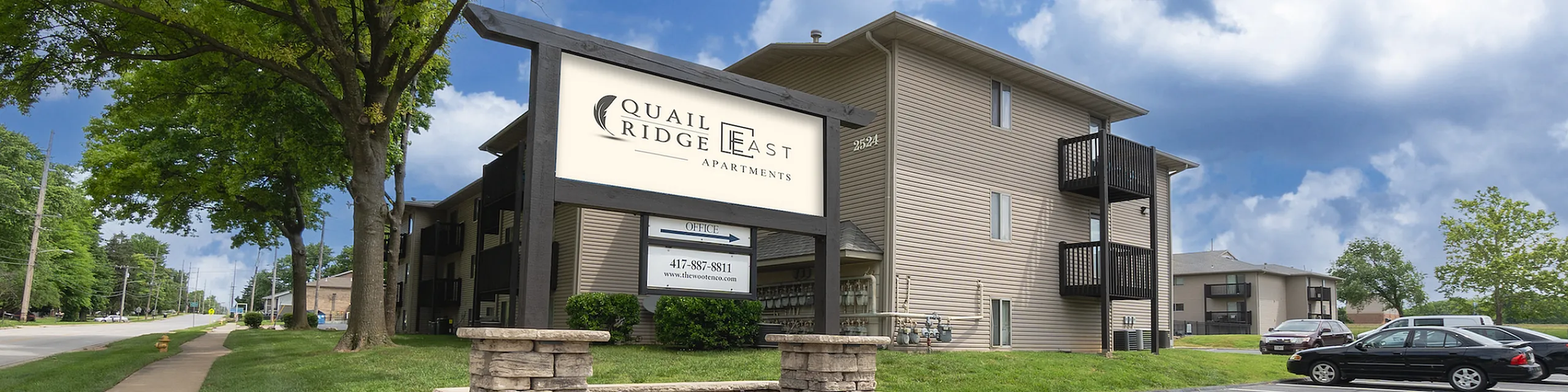 Sunny exterior view of Quail Ridge East Apartments with lush landscaping and ample parking, perfect for a serene living experience.