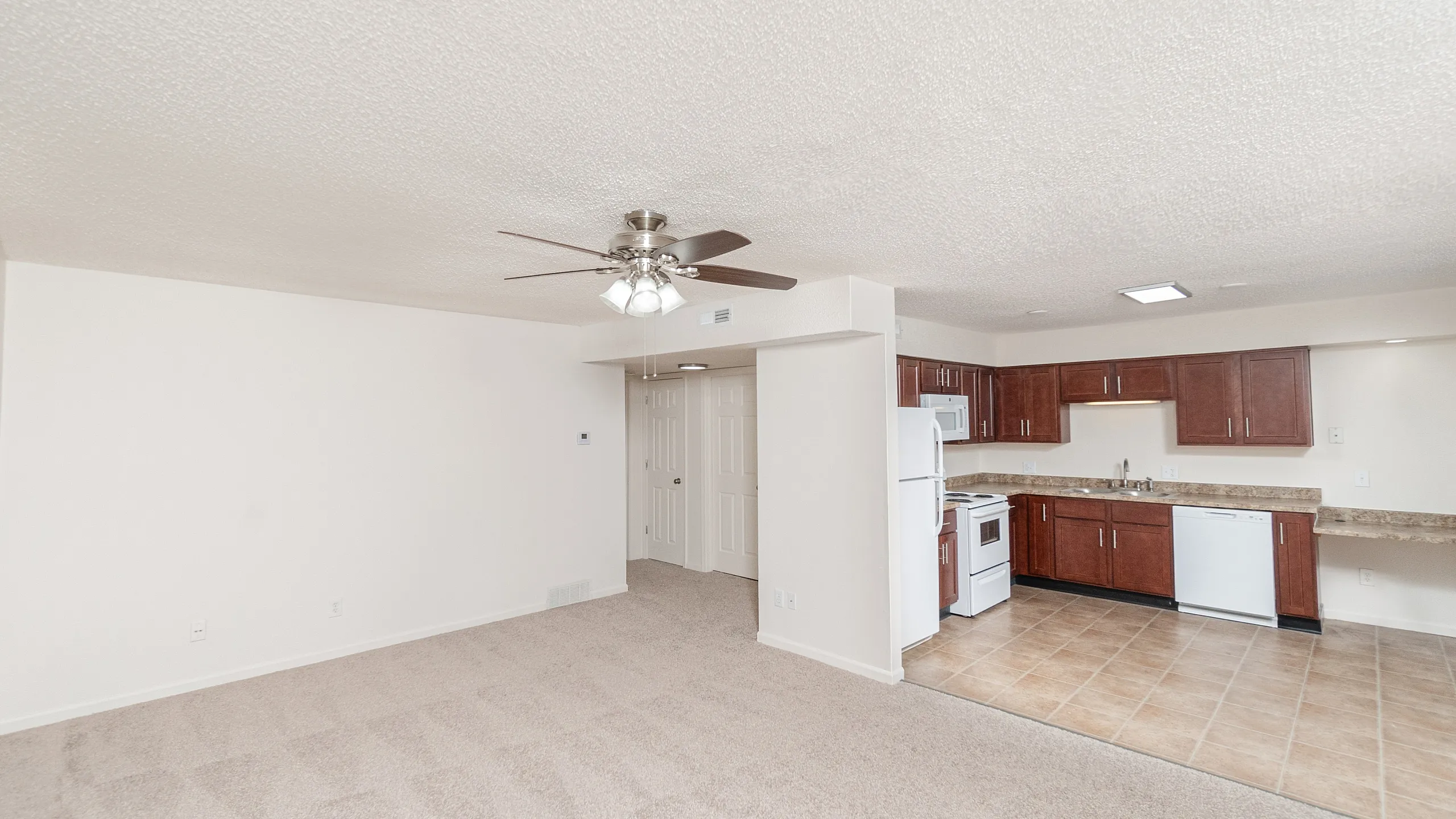 Modern open-plan living area featuring a spacious kitchen with rich wooden cabinetry, stylish appliances, and a cozy carpeted living space with a ceiling fan.