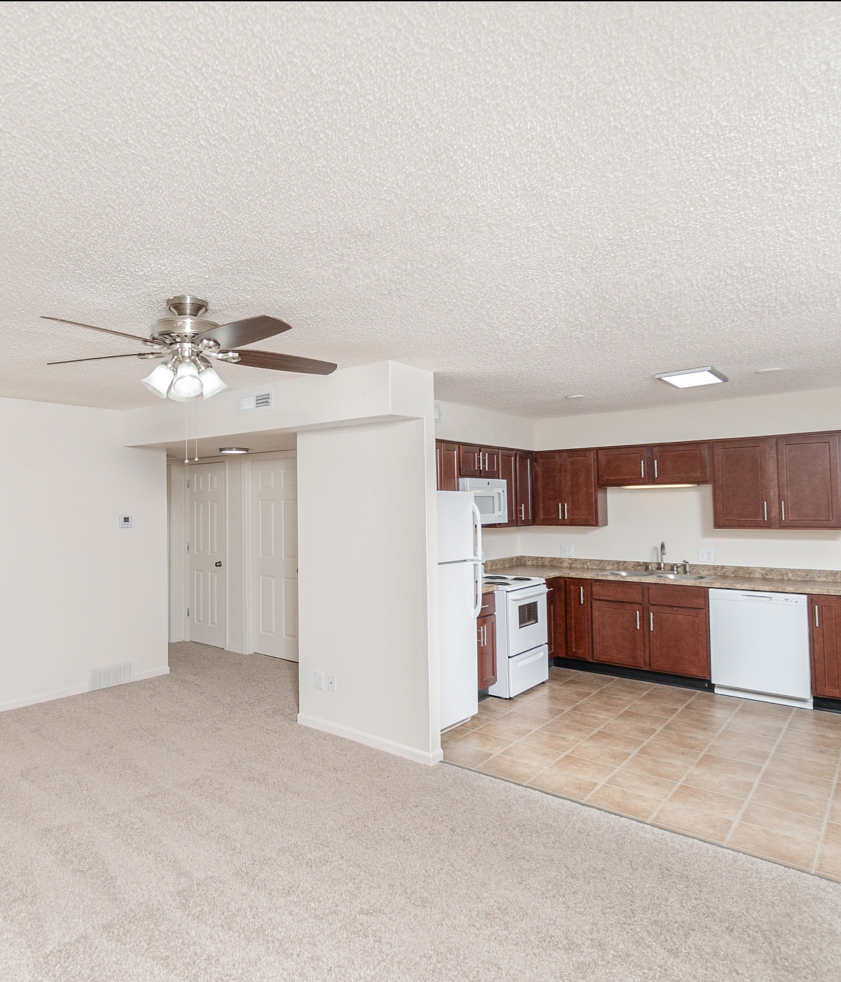 Bright and spacious open-plan living area with plush carpet, modern ceiling fan, and sleek kitchen featuring cherry wood cabinets and tiled flooring.