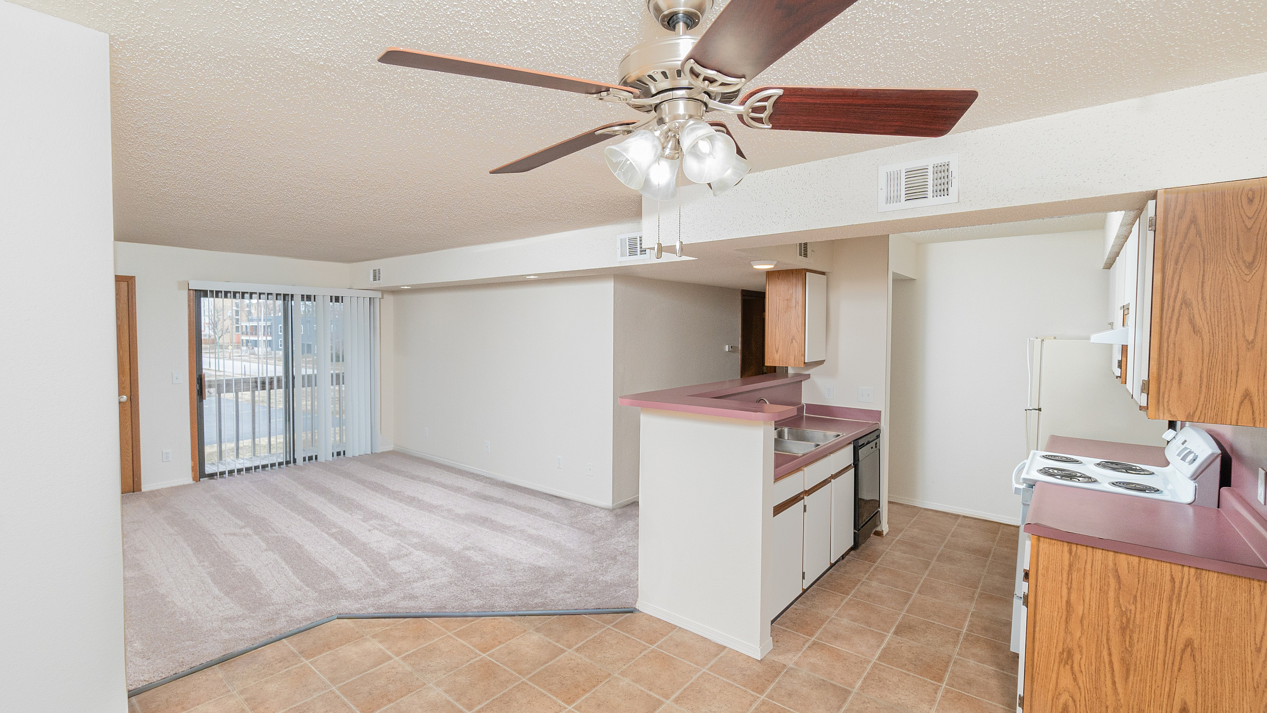 Open-concept TSH Apartments living space with carpeted living room, tile kitchen flooring, breakfast bar, wood cabinetry, ceiling fan with lights, and sliding glass door leading to patio.