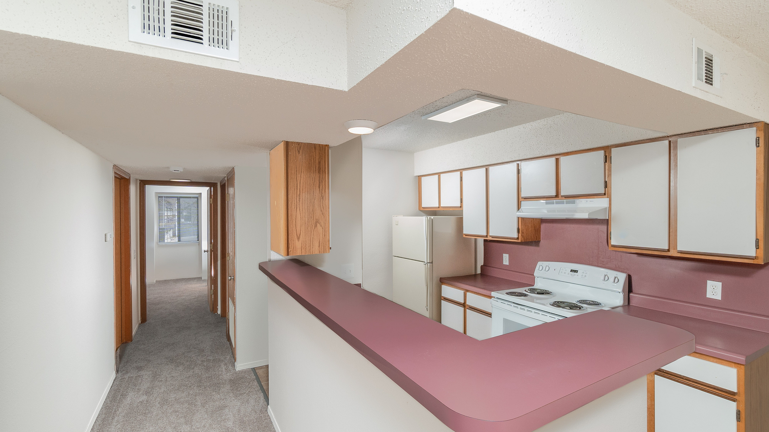 TSH Apartments kitchen with breakfast bar, pink countertops, white appliances, wood cabinetry, overhead lighting, and hallway leading to carpeted bedrooms.