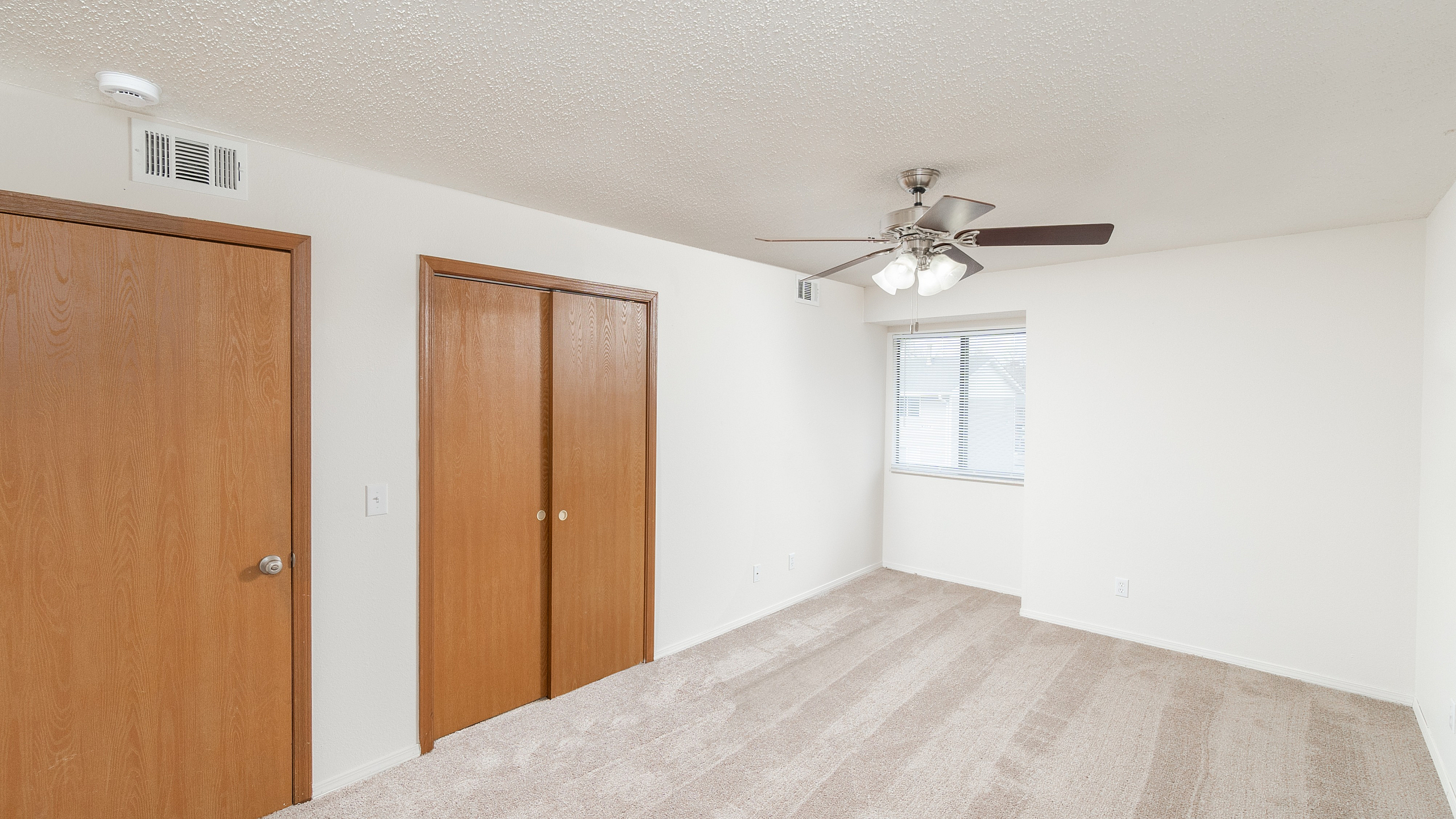 TSH Apartments bedroom with neutral carpet, white walls, wood closet doors, window with blinds, and ceiling fan with light fixture.