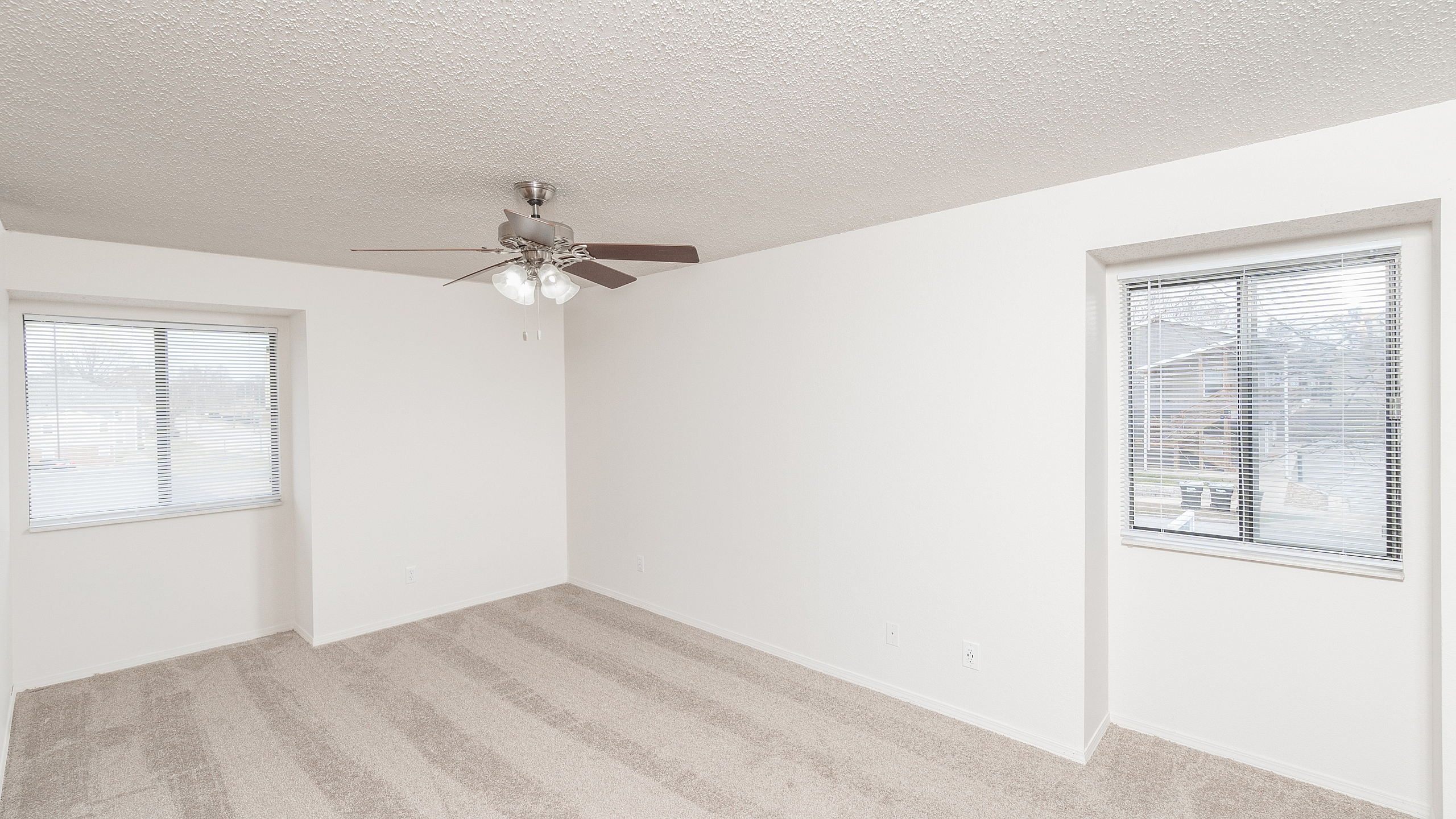 Spacious TSH Apartments bedroom with neutral carpet, white walls, two large windows with blinds, and ceiling fan with light fixture.