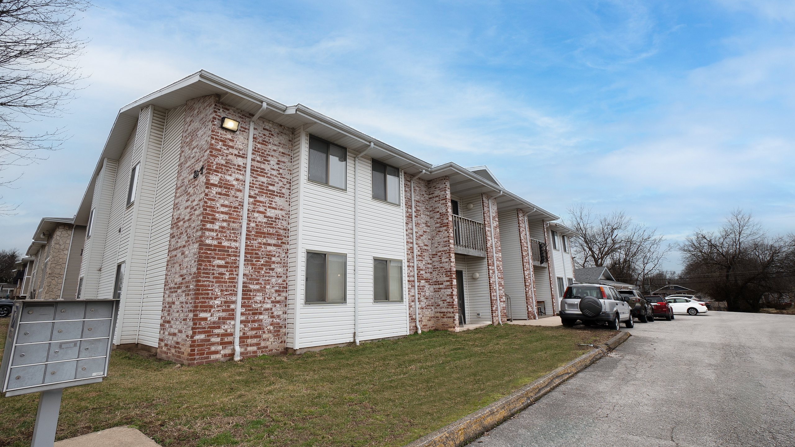 Exterior view of TSH Apartments featuring two-story brick and siding buildings, on-site parking, balconies, and community mailbox area.