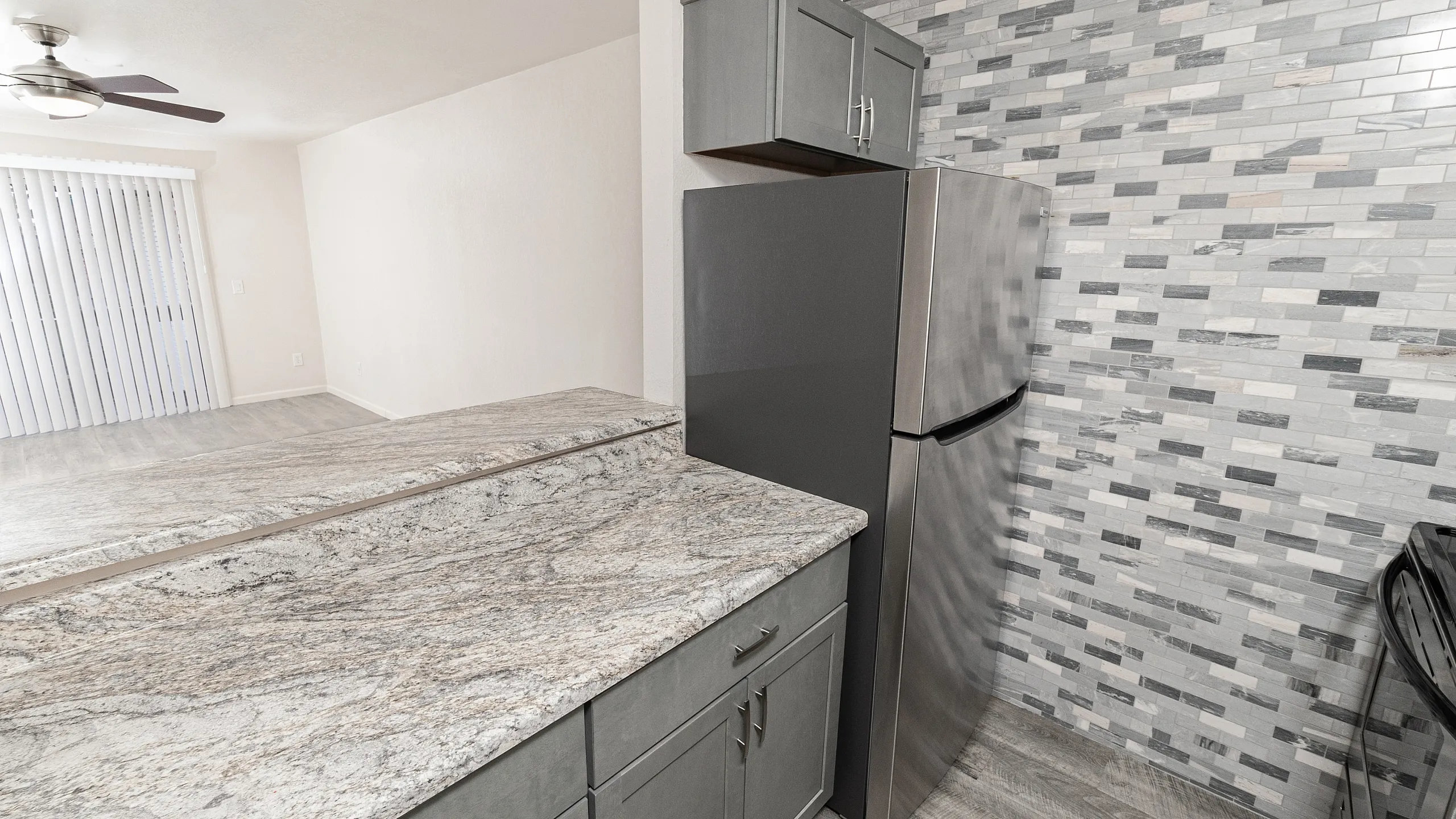 Modern kitchen showcasing sleek stainless steel refrigerator, elegant gray cabinets, and a stylish mosaic tile backsplash, leading to an open living space with natural light.