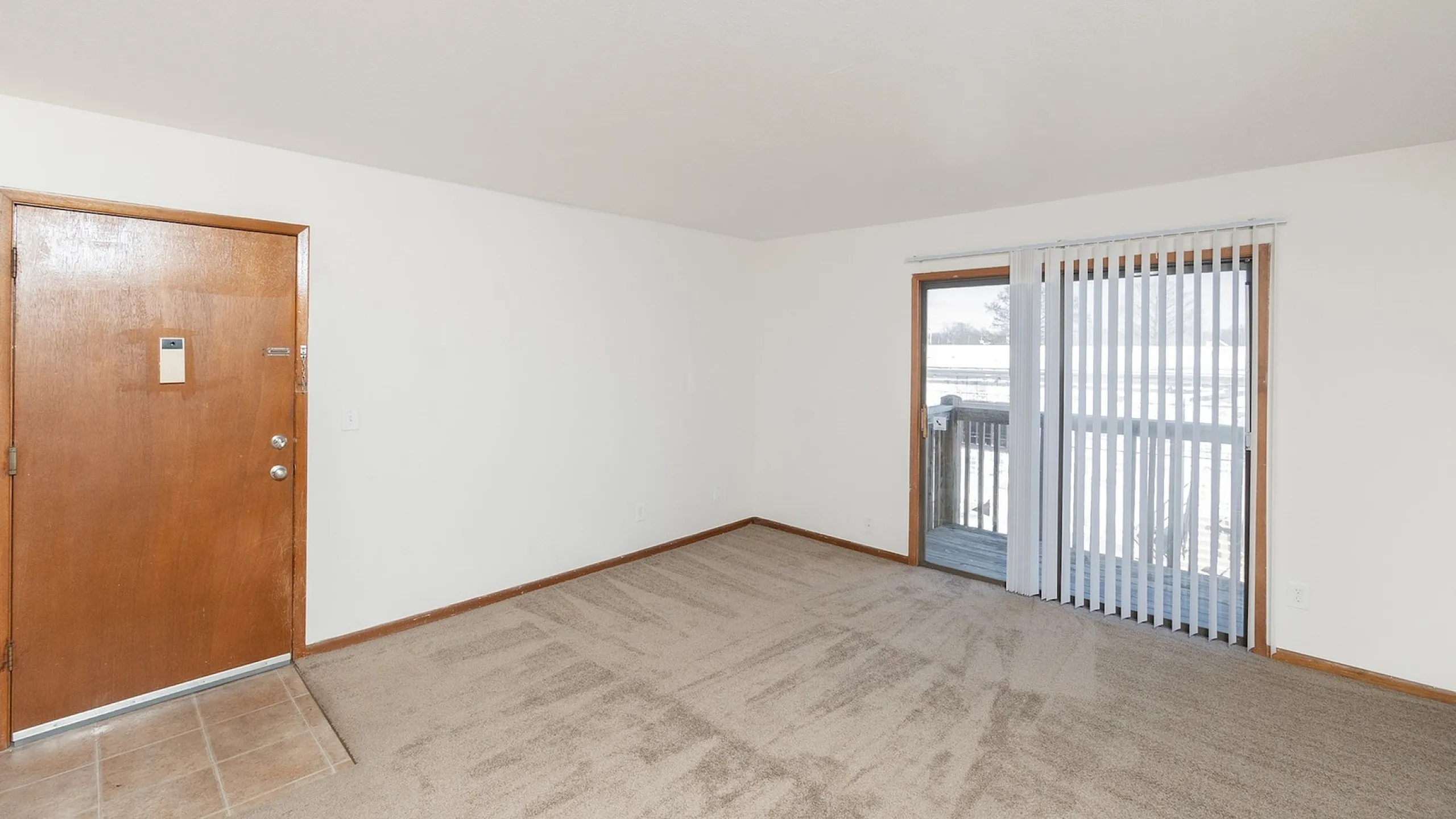 Bright living room with plush carpet, wooden trim, and glass door leading to a sunlit balcony, perfect for relaxation.