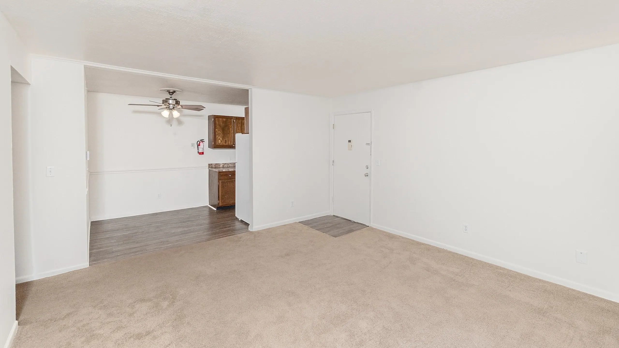 Bright open living space with neutral walls, featuring a cozy carpeted area leading into a sleek wood-floored kitchen with stylish lighting.