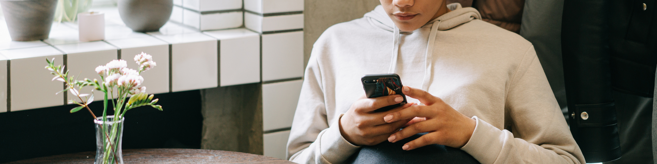 Person sitting at a table holding a smartphone near a window with plants and soft natural light.