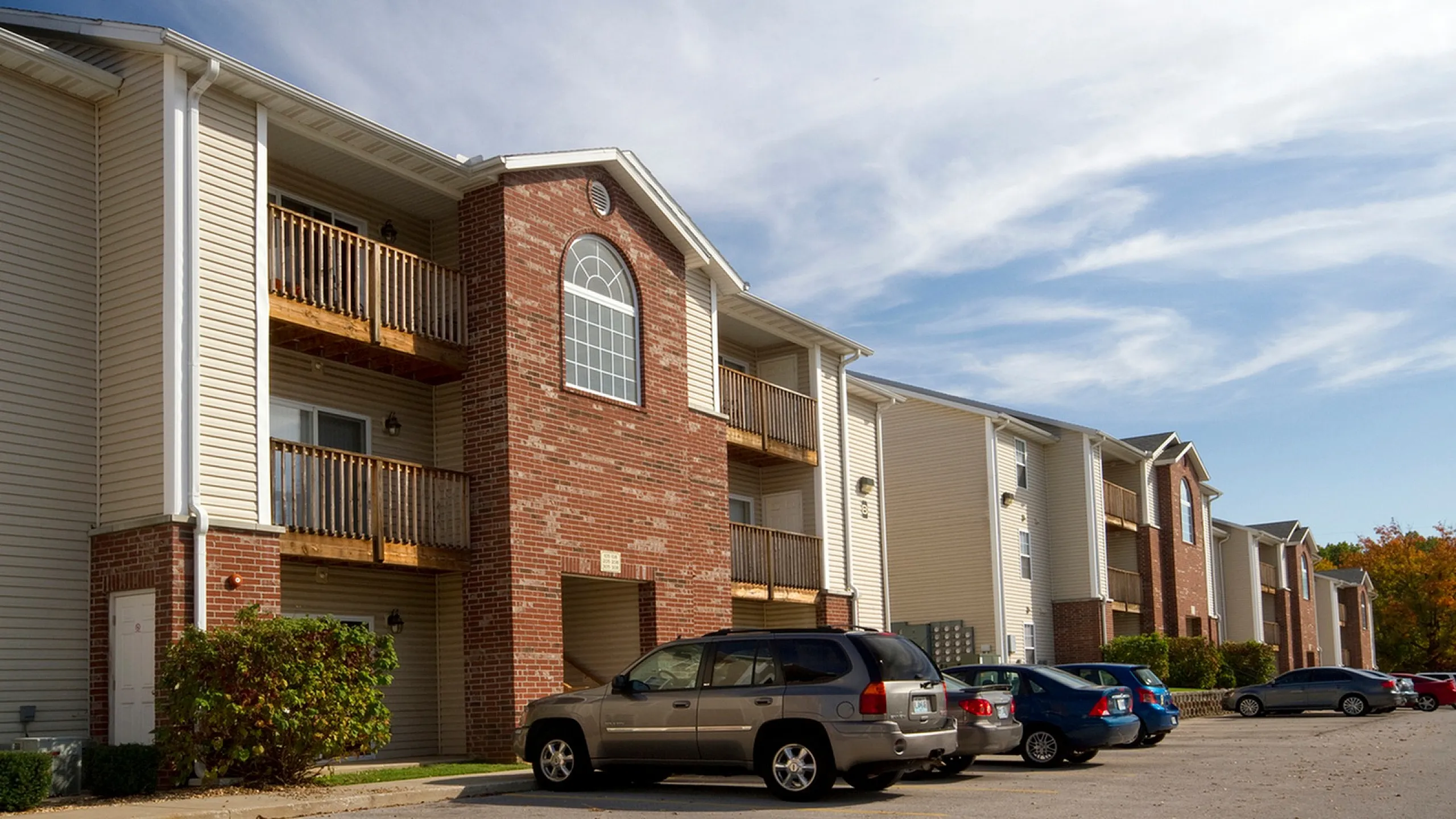 Three-story apartment community with red brick and beige siding, featuring private balconies, large windows, and a surface parking lot with resident vehicles under a clear blue sky.