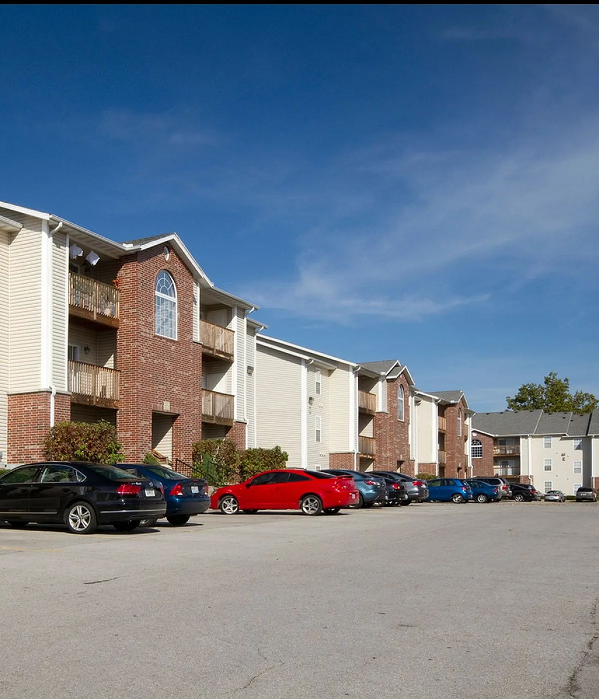 Three-story apartment community with red brick and beige siding, featuring private balconies, large windows, and a surface parking lot with resident vehicles under a clear blue sky.