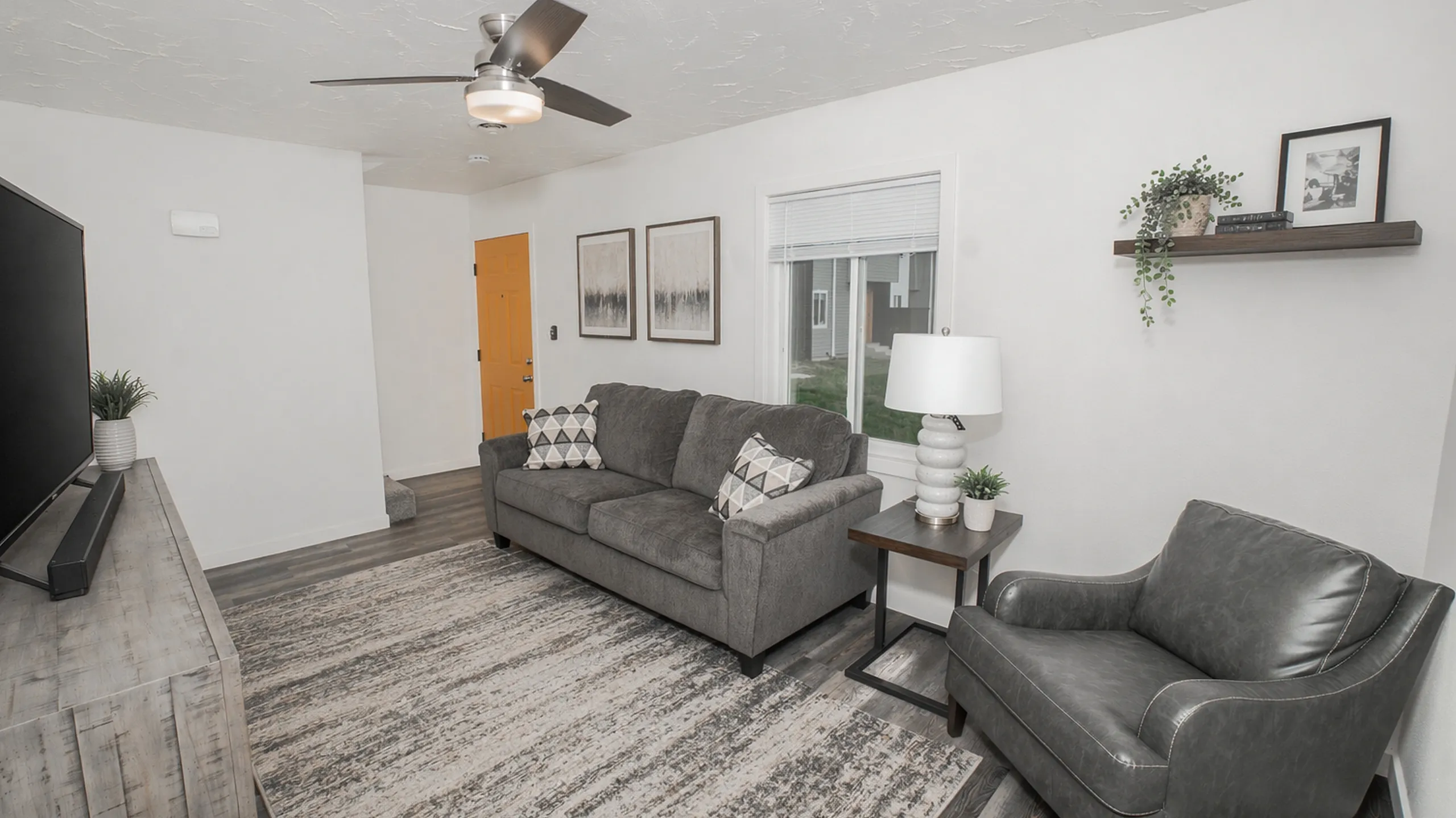 Cozy living room with gray sofa, accent chair, large area rug, wood TV console, side table with lamp, wall art, and window providing natural light near the entry door.