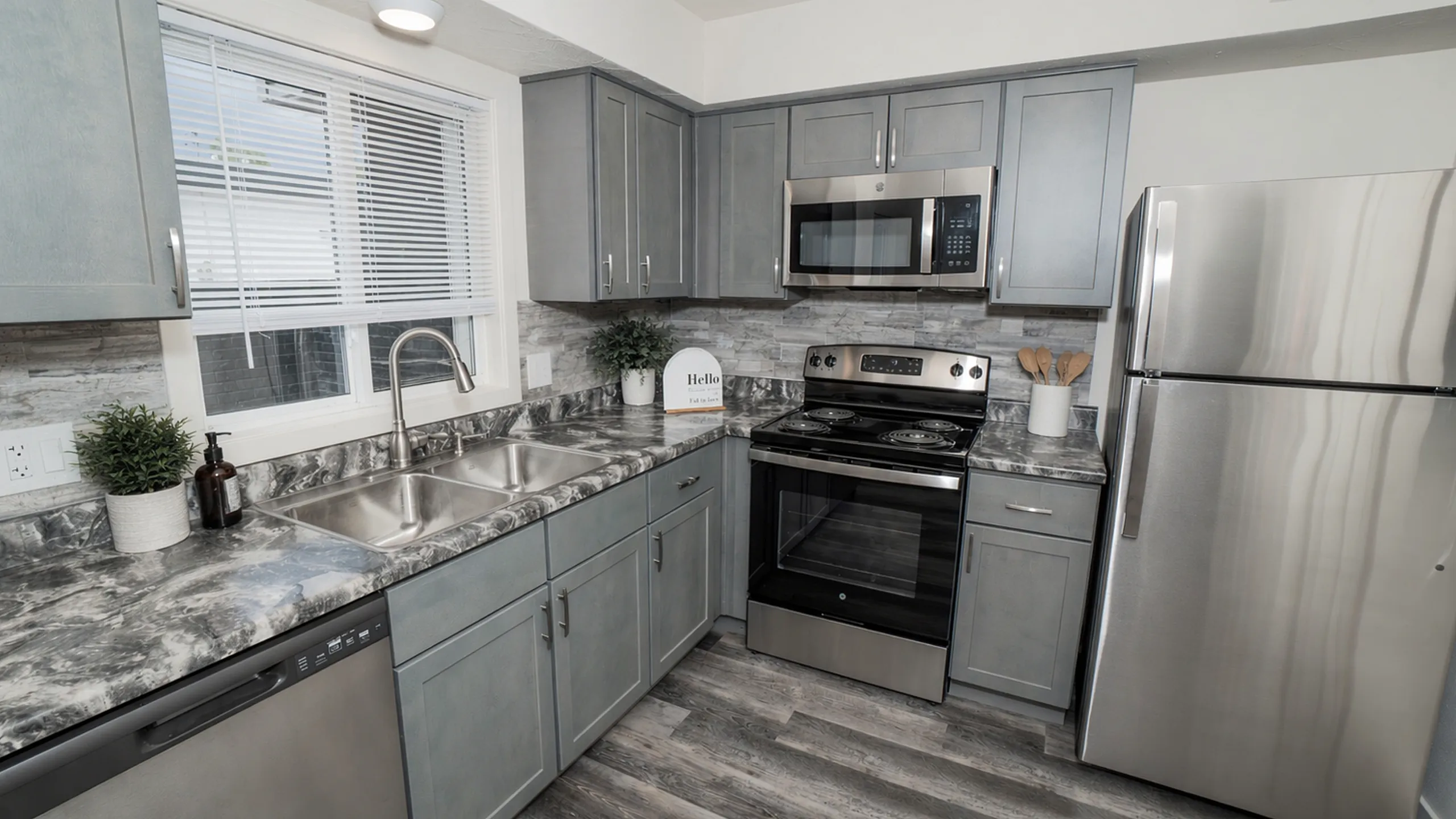 Updated kitchen with gray cabinets, stainless steel appliances, double sink, modern faucet, marble style countertops, tile backsplash, and wood style flooring.