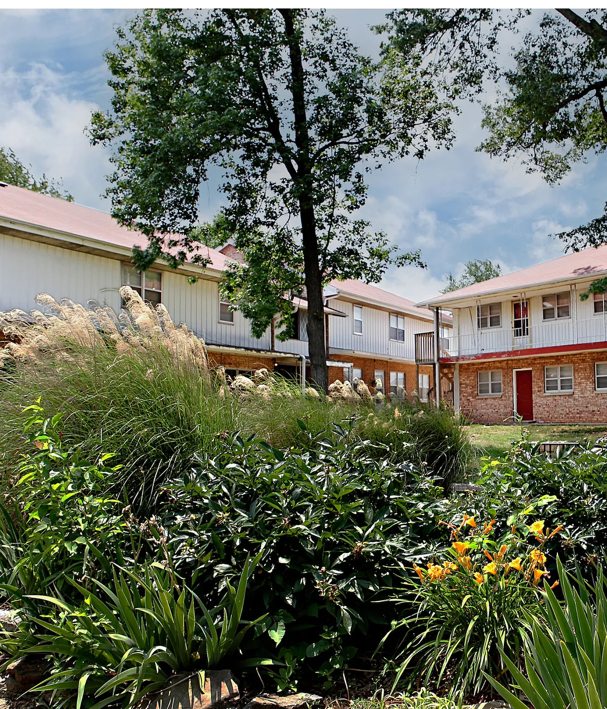 Charming apartment complex with brick and siding exterior nestled among lush greenery and vibrant flowers, offering a serene and inviting atmosphere.