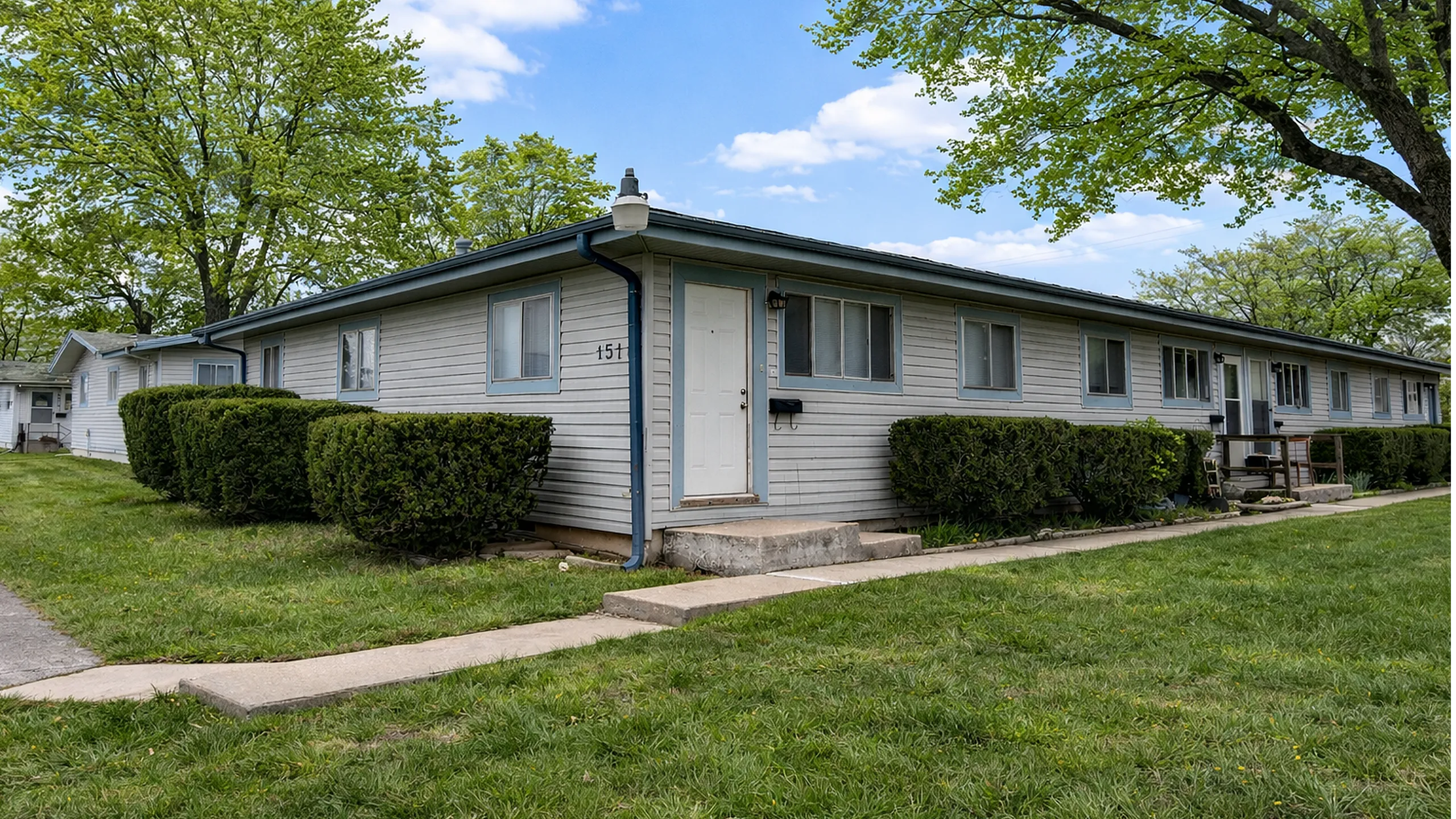 Charming single-story quadplex building with lush greenery and welcoming entrance, set against a vibrant blue sky.