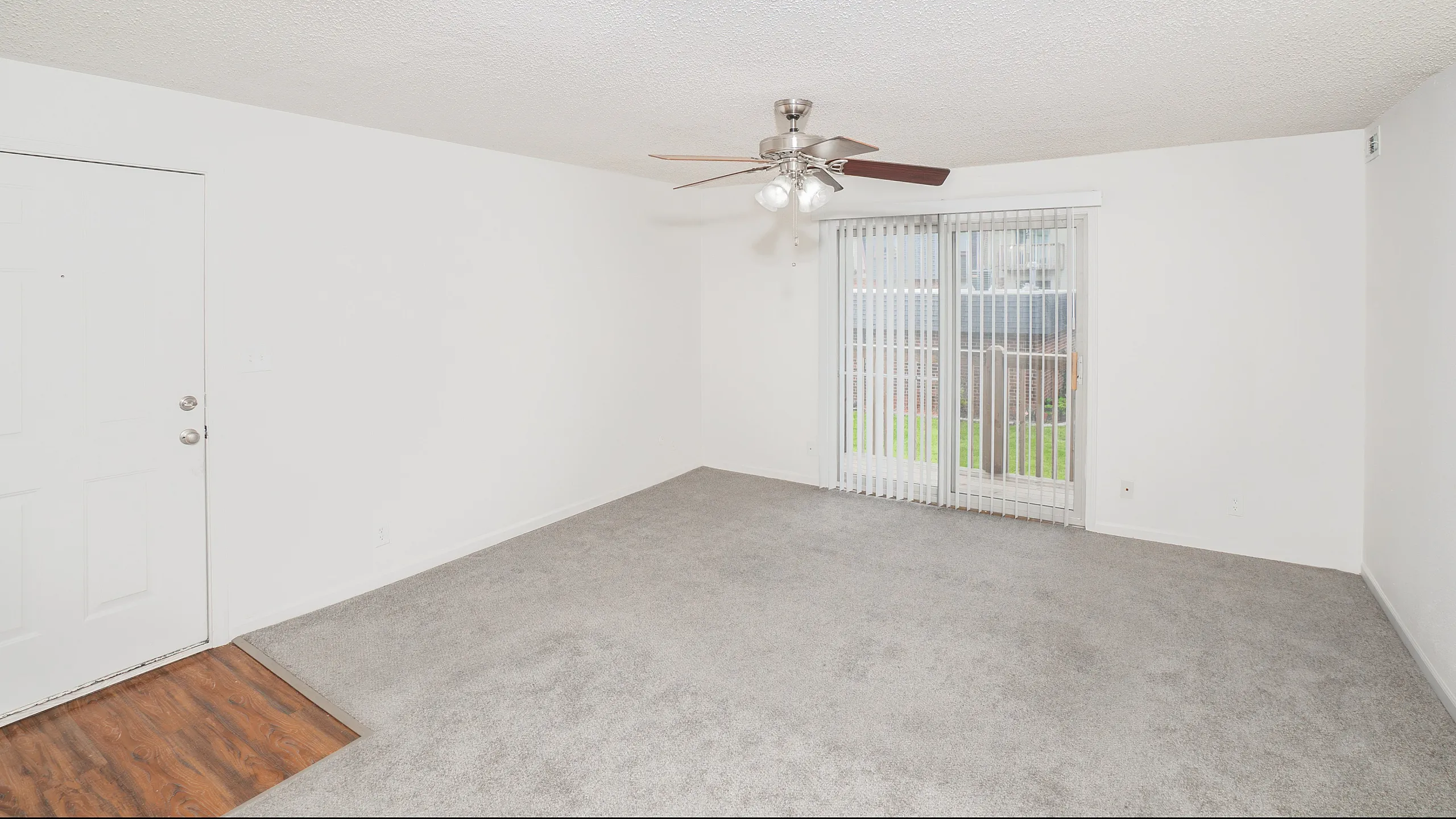 Bright and inviting living room with plush carpet, elegant ceiling fan, and sliding glass doors leading to a serene outdoor view.