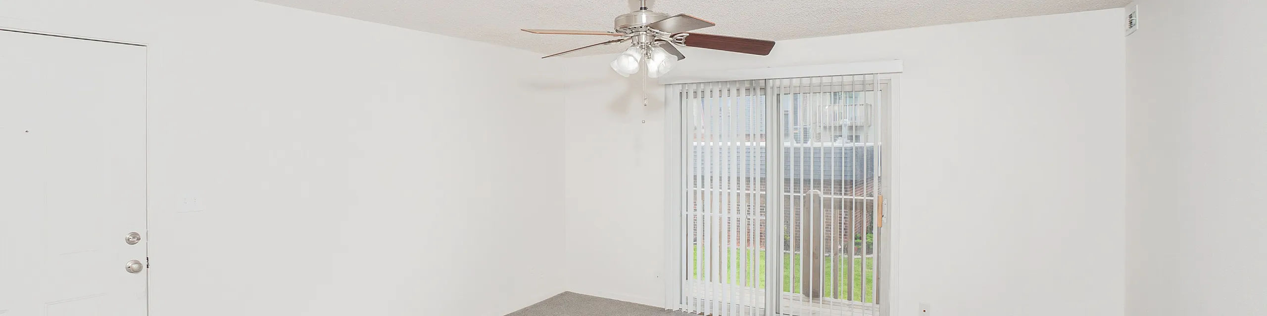 Bright and airy living room with plush carpet, ceiling fan, and sliding glass doors leading to a serene outdoor space.