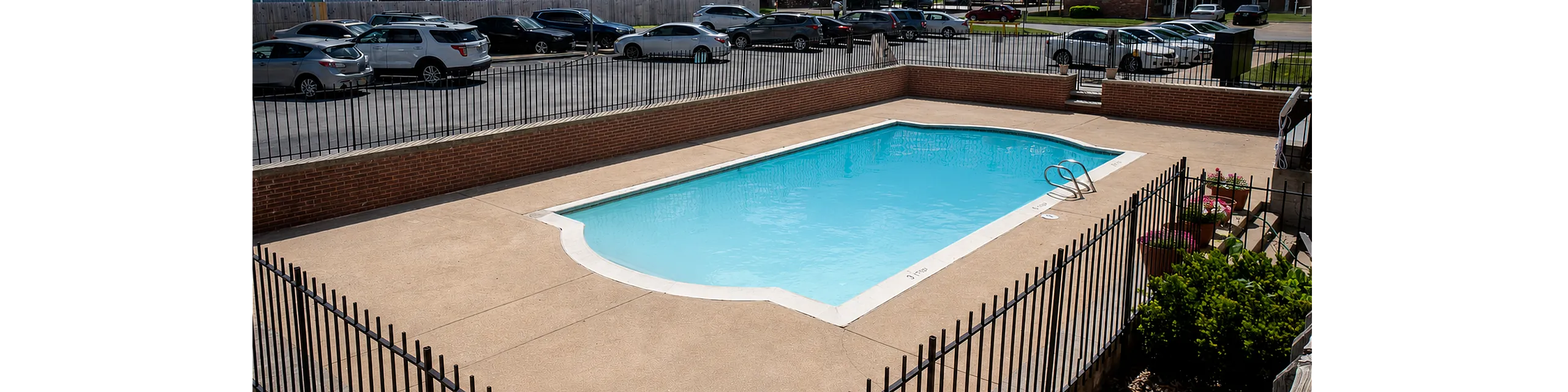 Inviting outdoor swimming pool surrounded by secure black fencing, set in a vibrant apartment community with ample parking and lush greenery.